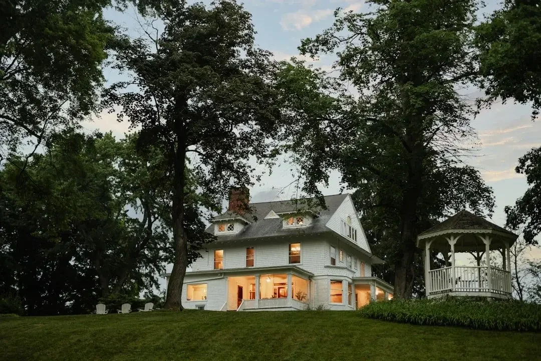 A large white Victorian-style house with multiple stories, lit from inside, surrounded by large trees. A small white gazebo with a railing is on a grassy hill nearby.