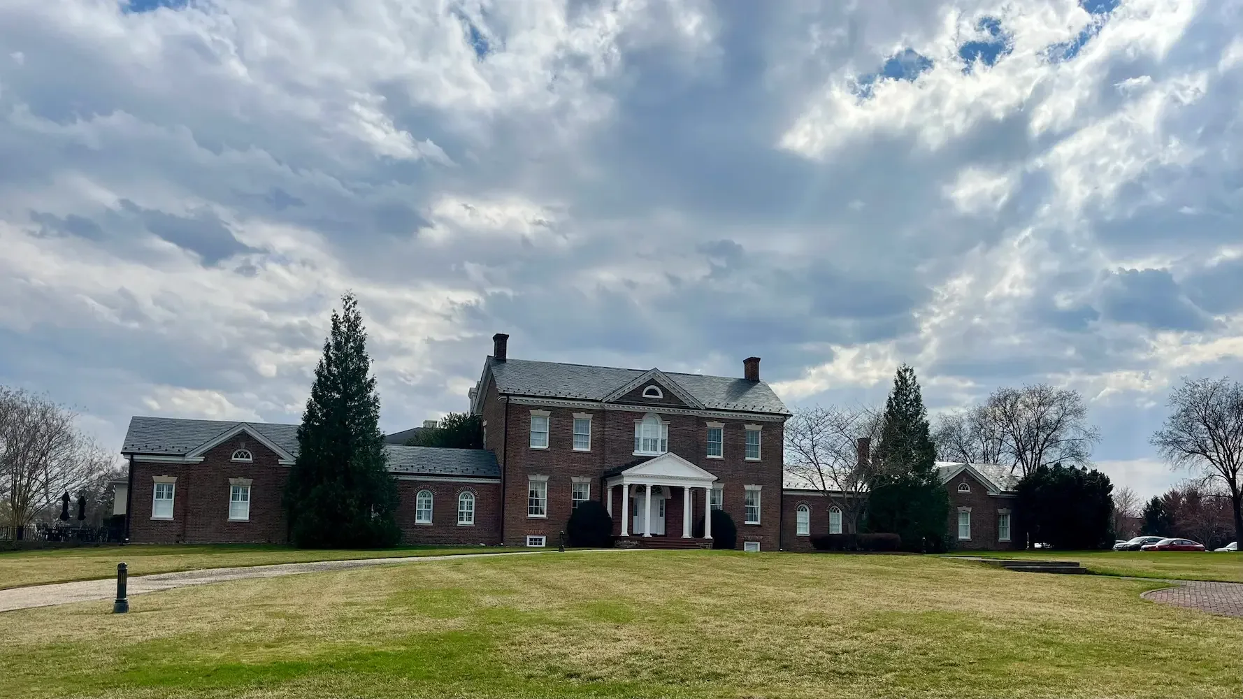 Large historic brick house with white columns and a front porch, surrounded by a well-maintained lawn and trees under a cloudy sky.