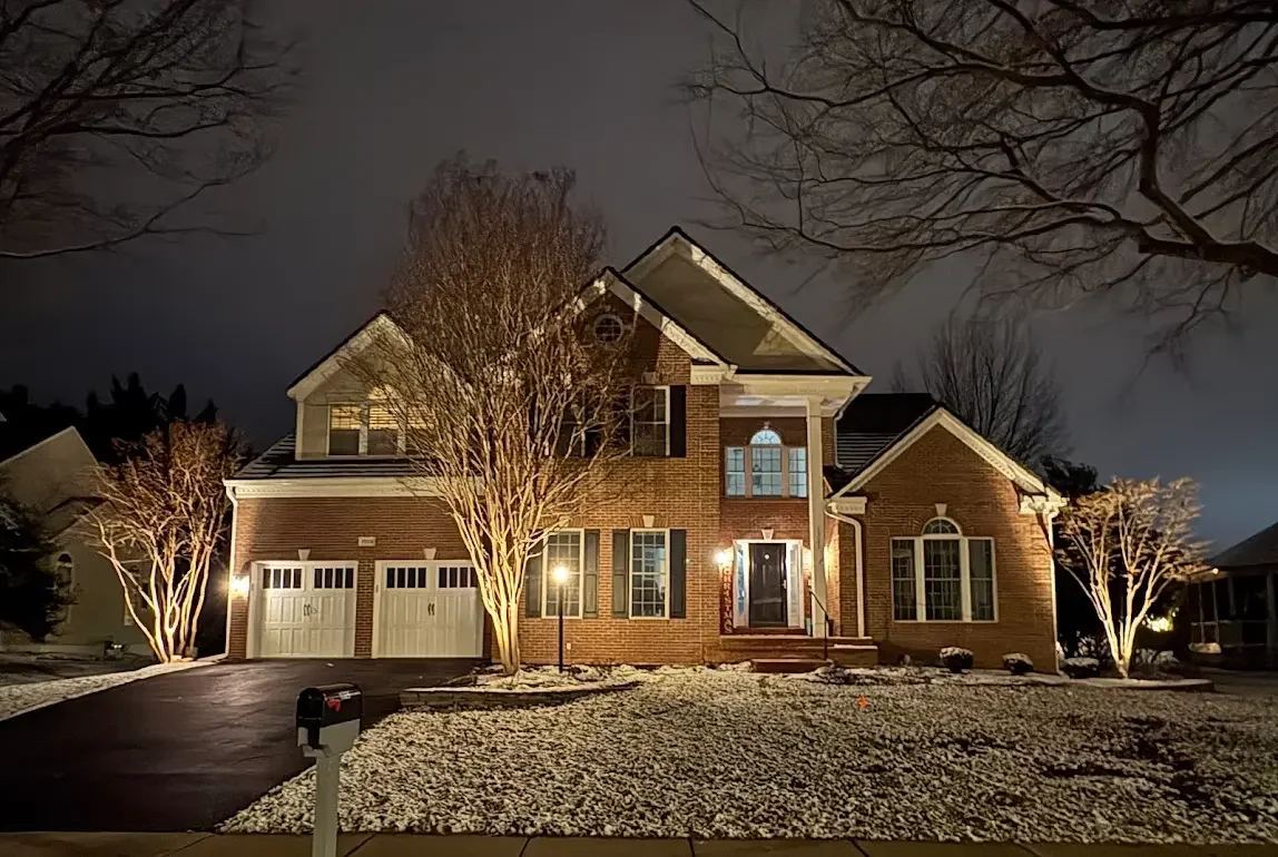 A two-story brick house at night with exterior lights on, snow on the ground and leafless trees in the front yard.