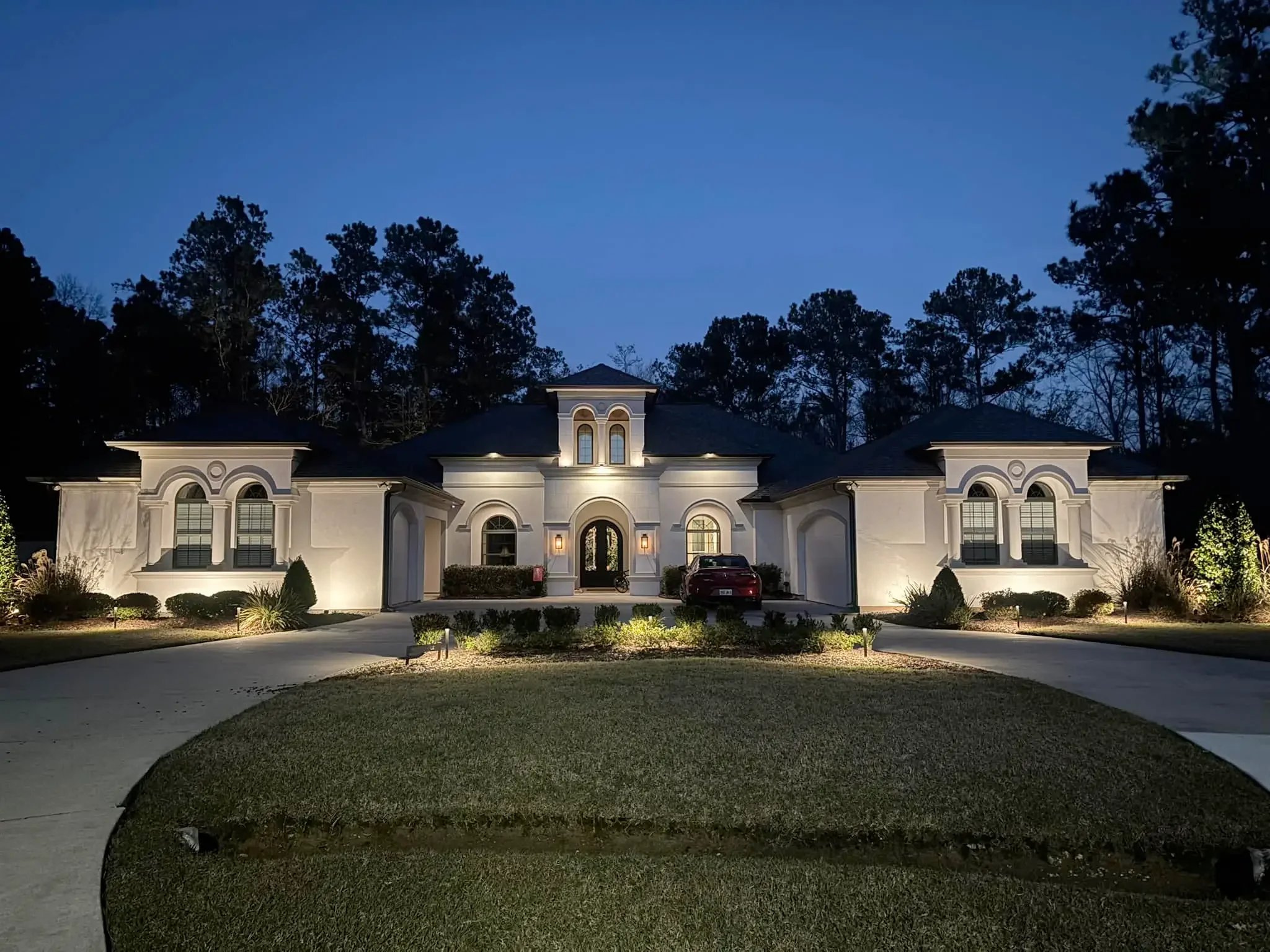 Large, white, two-story house illuminated at night with trees in the background, a curved driveway, and a well-maintained lawn in the front.