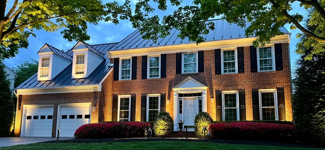 A large brick two-story house illuminated at dusk, with black shutters and a green lawn.