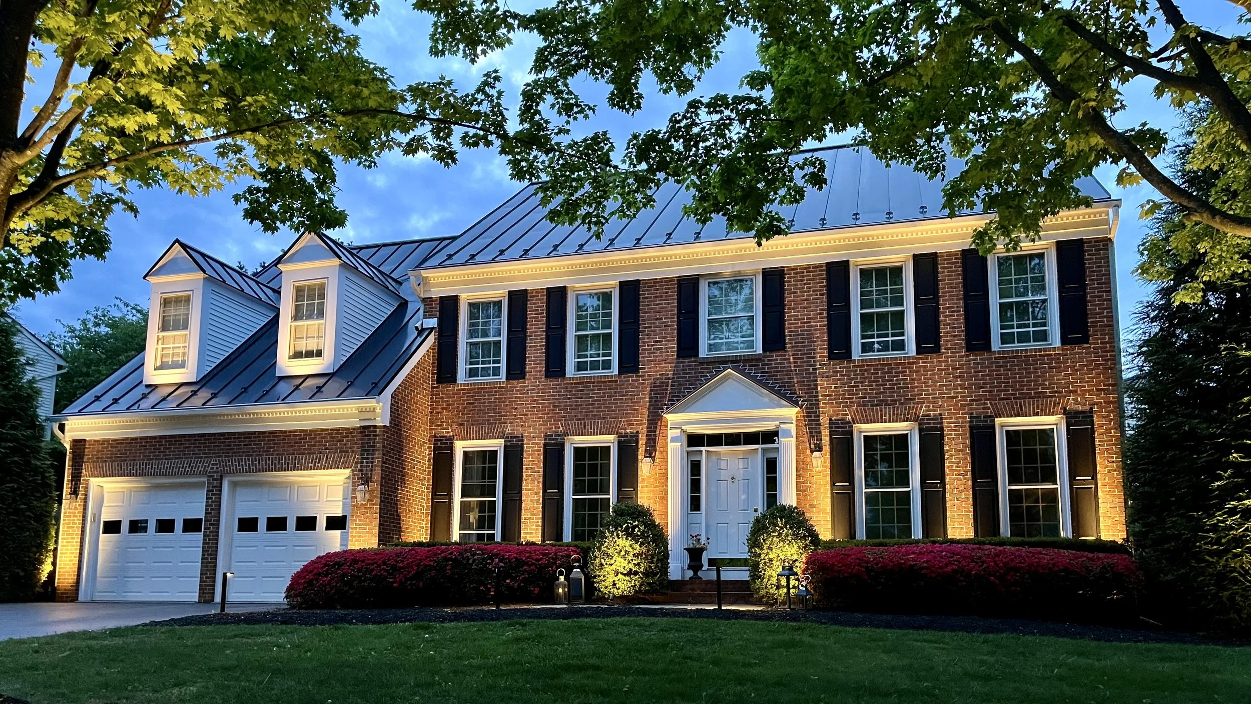 A large brick house in Leesburg, VA with black shutters and a metal roof, illuminated by exterior lights at dusk, with well-maintained landscaping including bushes and trees.