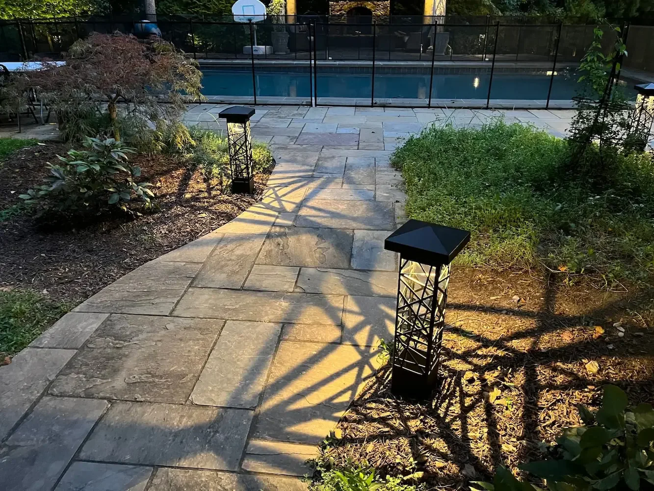 A backyard with a paved stone pathway leading to a swimming pool, flanked by garden beds with plants and shrubs, illuminated by warm exterior bollard lighting.