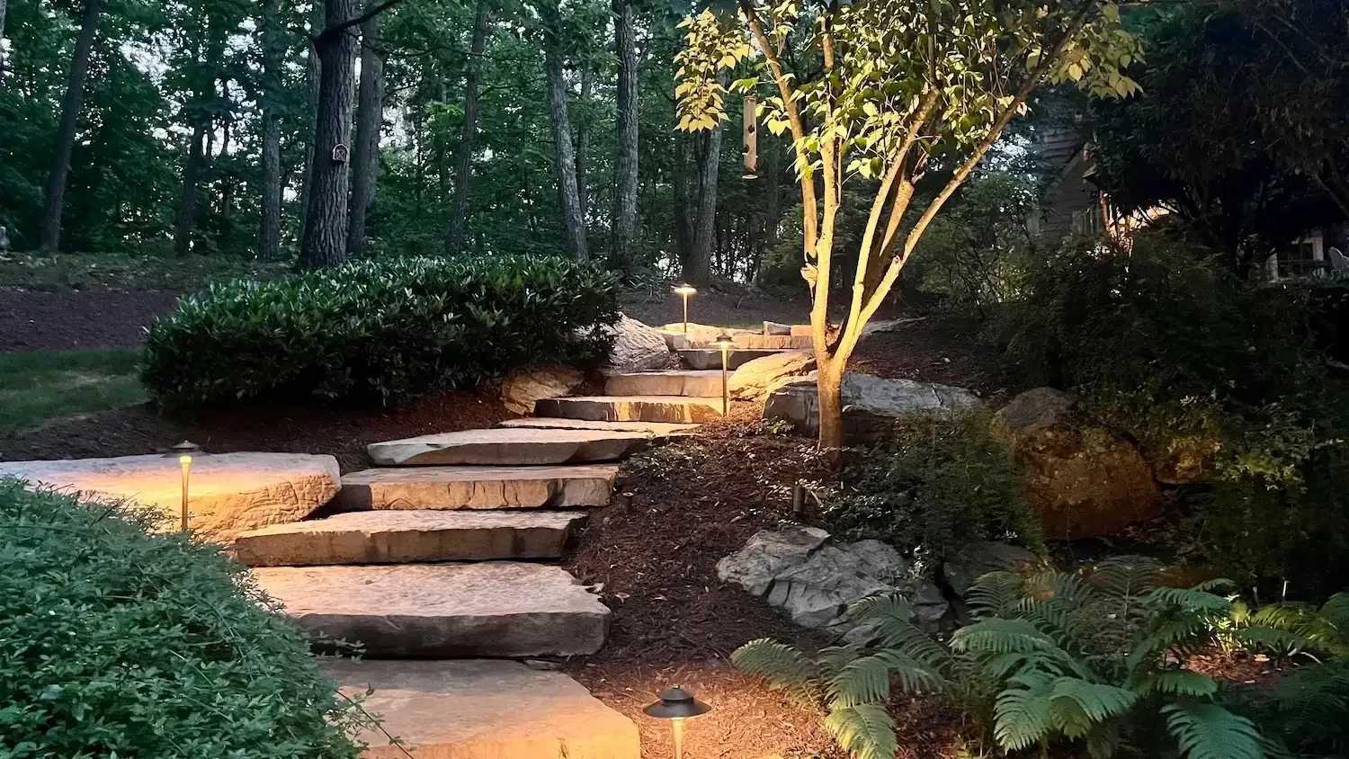 A stone pathway with steps leading uphill in a garden, illuminated by small landscape lights at dusk or evening, surrounded by trees, bushes, and large rocks.