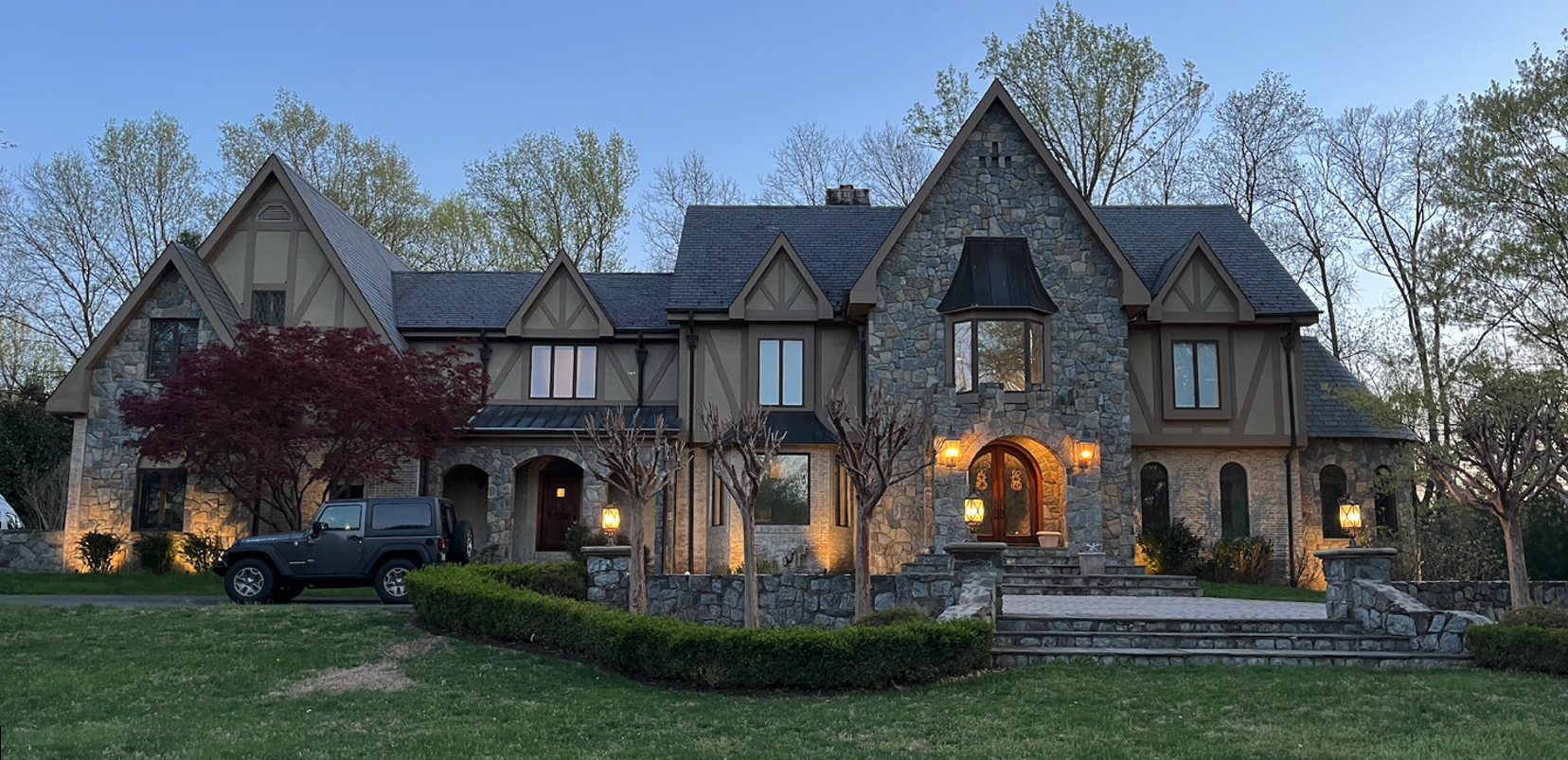 Large stone and wood Tudor-style house with triangular roofs, surrounded by trees and well-maintained lawn, with an SUV parked in driveway.