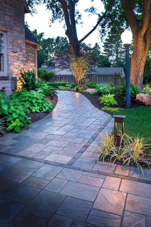 A landscaped backyard walkway made of gray paving stones curves past a stone house, surrounded by lush green plants and trees, with outdoor lighting.