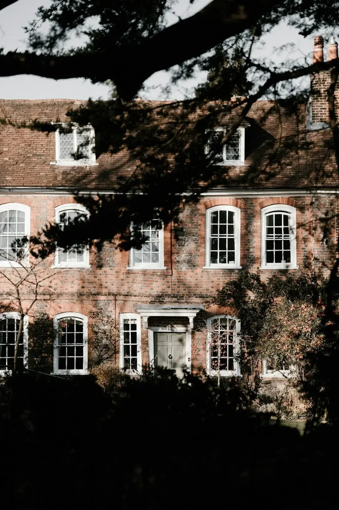 A multi-story brick house with white-framed windows, a green front door with a small overhang, and greenery in the yard, partially obscured by tree branches.