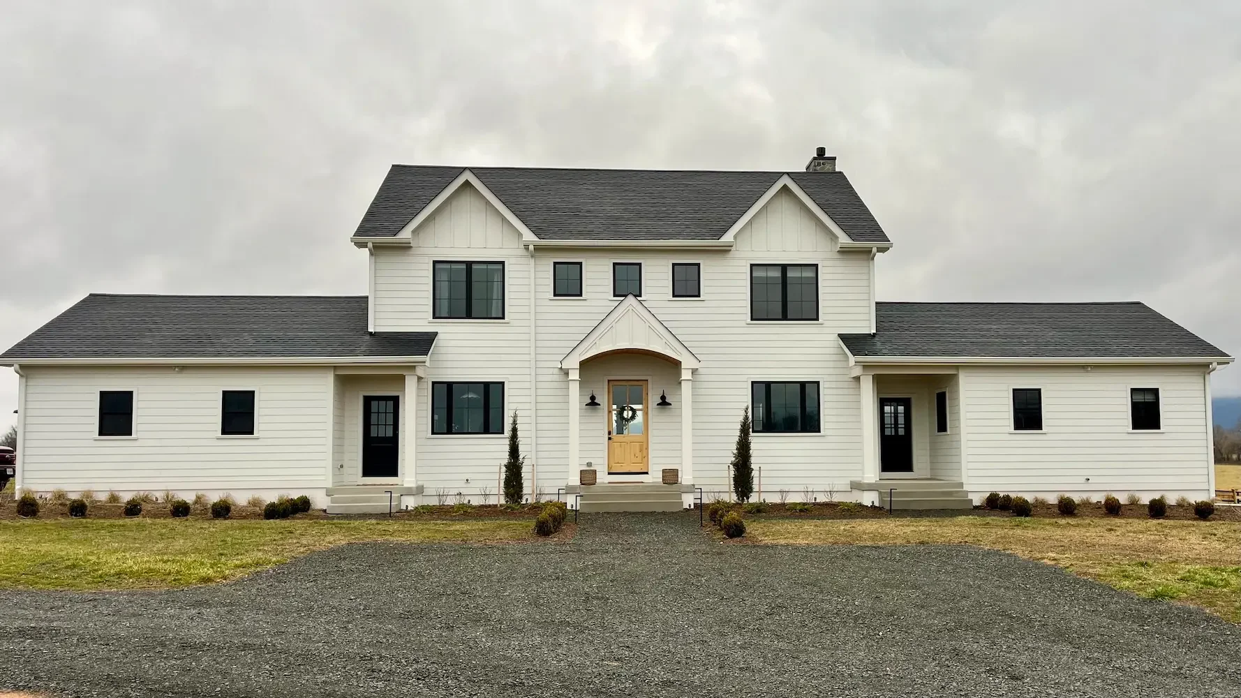 A large white two-story house with black roof shingles, front porch, and multiple windows, situated on a gravel driveway with sparse landscaping.