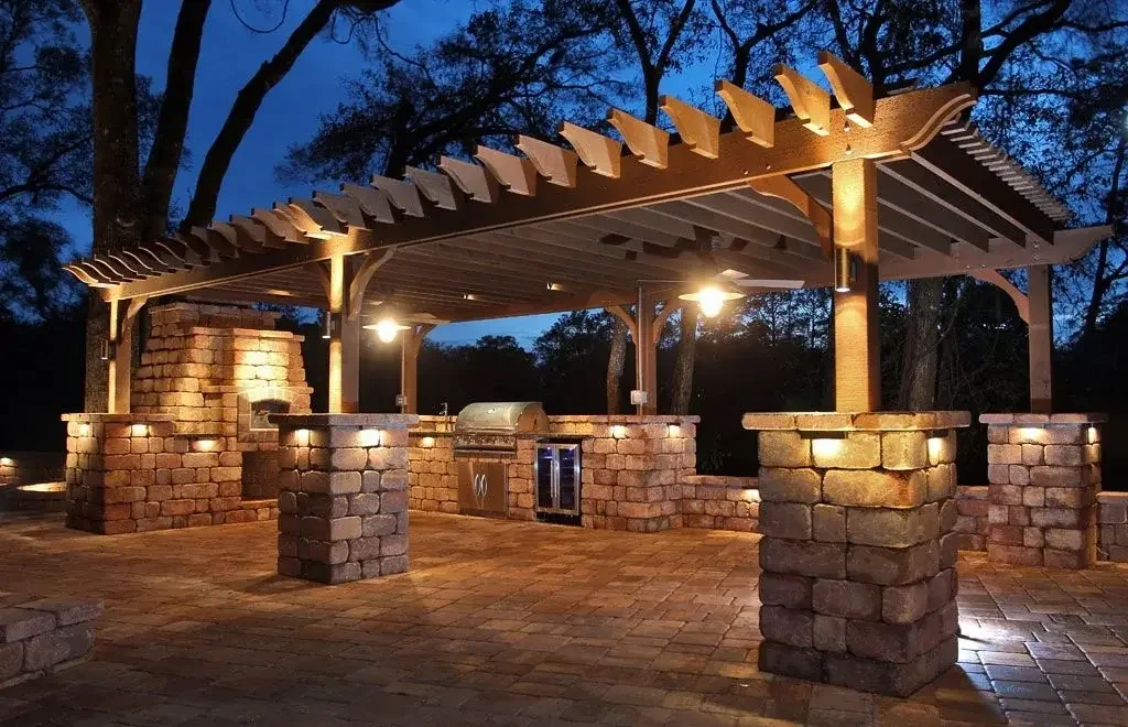 Outdoor brick outdoor kitchen with a roof and built-in grill, lit by ceiling lights, in a wooded area at dusk.