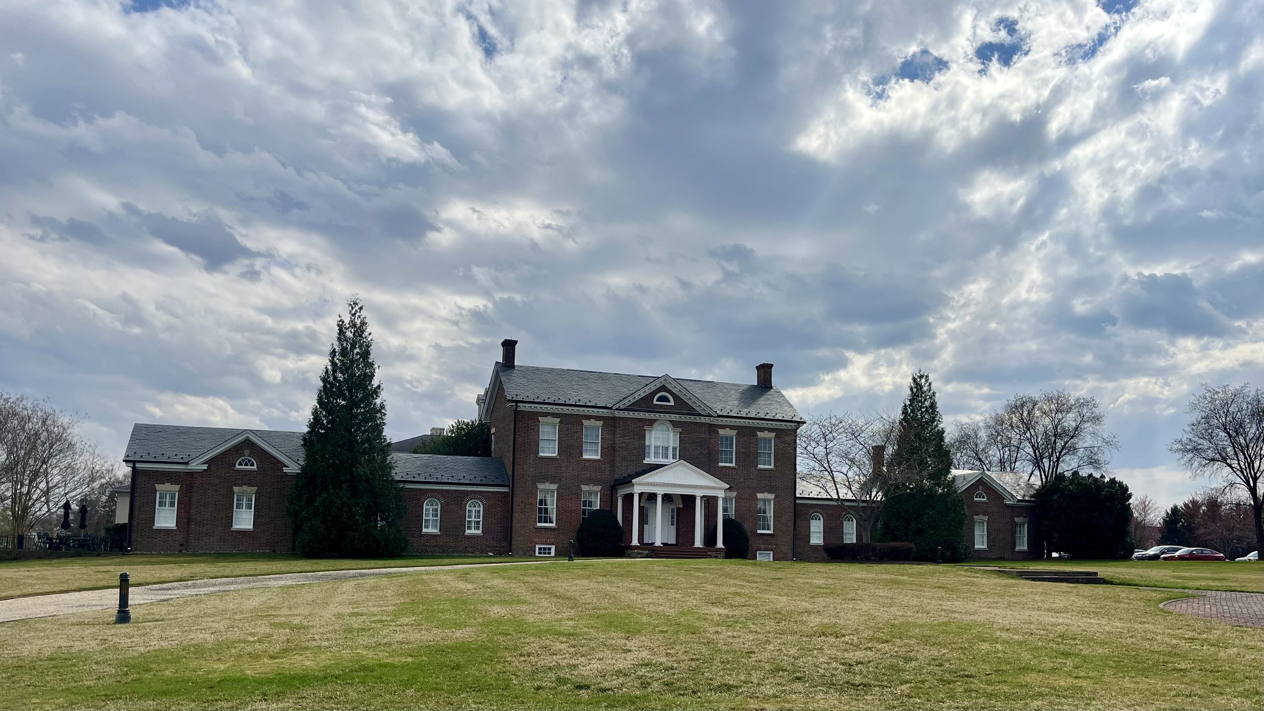 Large brick mansion with white columns and a spacious lawn under a partly cloudy sky.