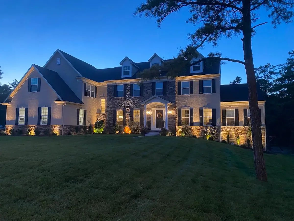 Large modern house with stone and siding exterior, front porch, multiple windows, landscaped yard, trees, and outdoor lighting, during twilight.
