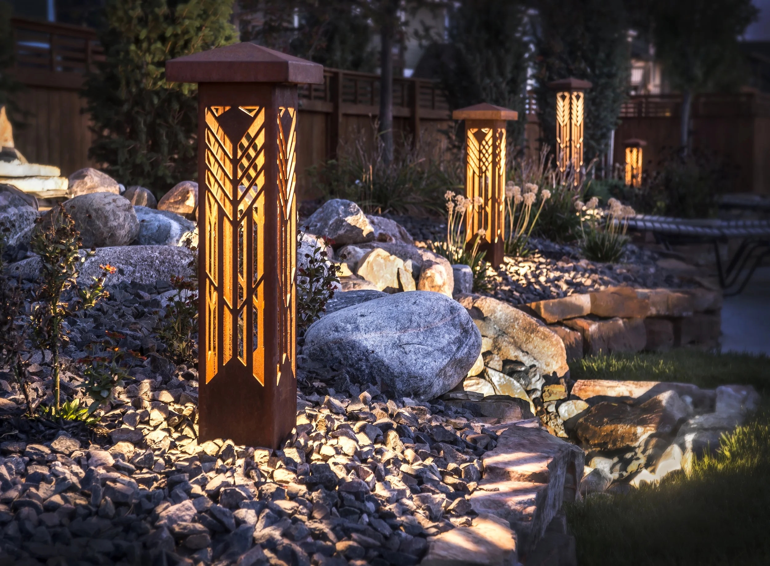 Outdoor pathway illuminated by decorative bollard lights, with rocks, plants, and a wooden fence in the background during evening.