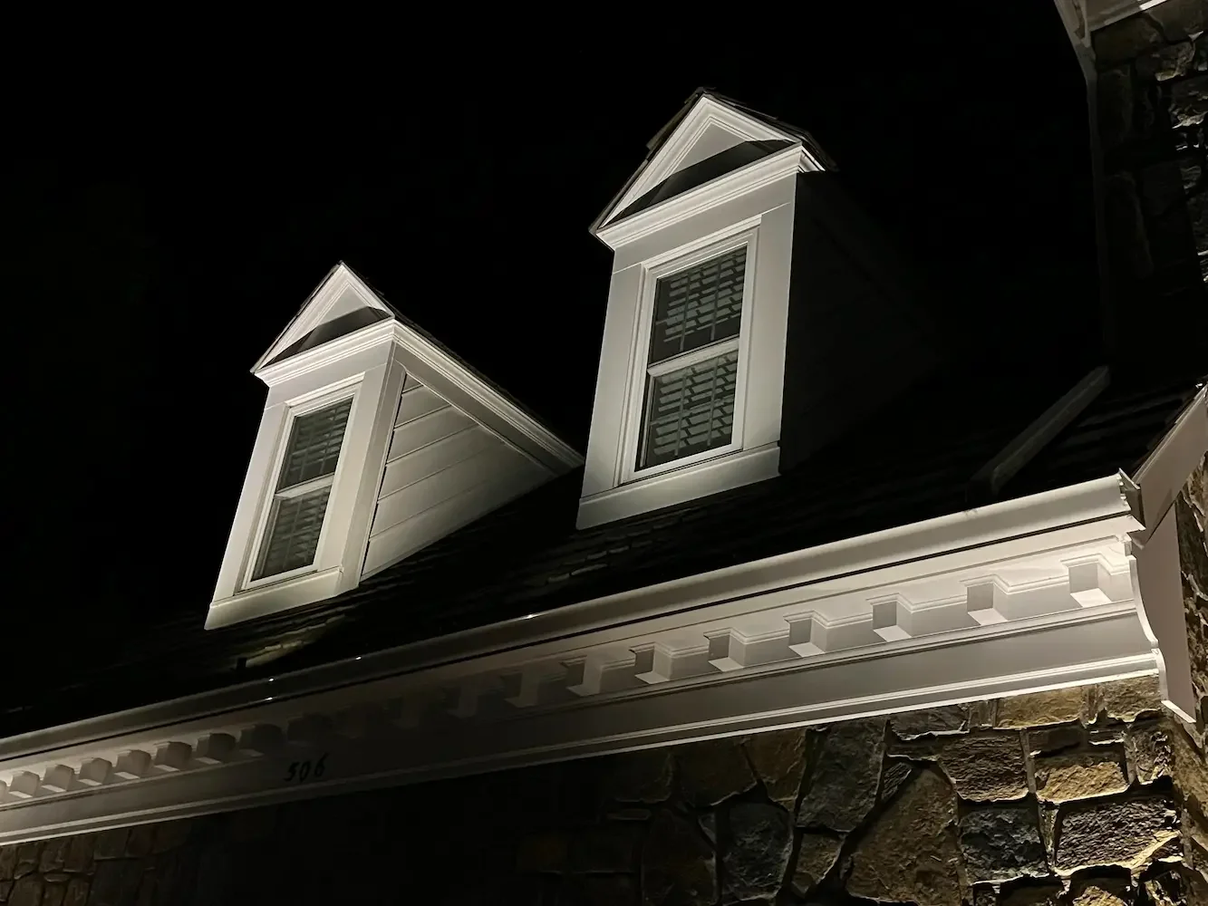 Two white dormer windows with black shutters on a house roof at night, with white decorative trim.