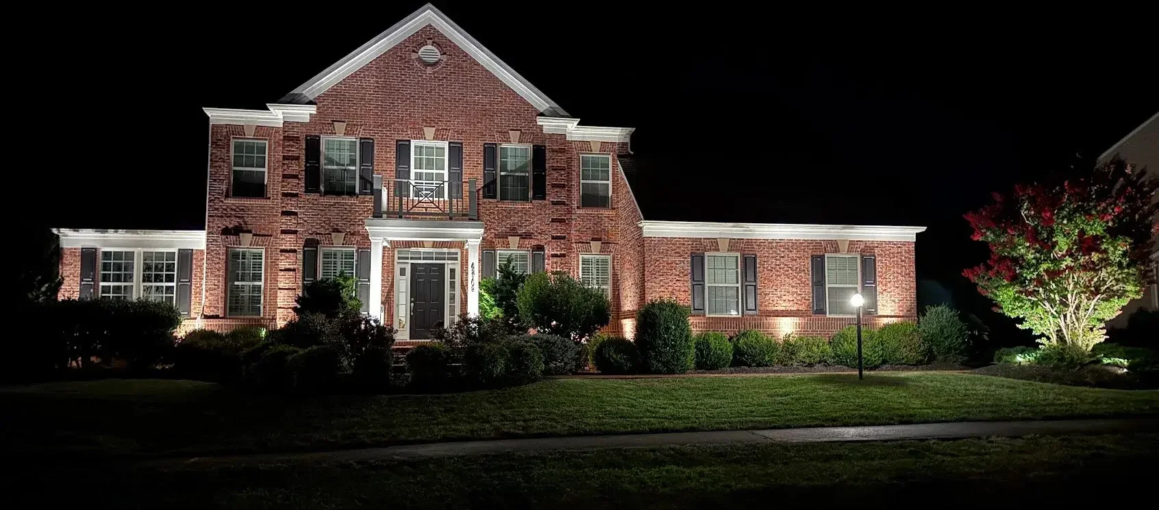 A large brick house illuminated at night with exterior lights, surrounded by well-maintained bushes, trees, and a lawn.