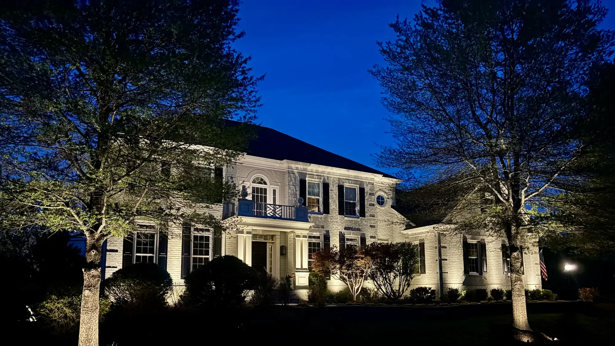 A large white house with black shutters, illuminated with architectural lighting, surrounded by uplit trees with some leaves, and a well-maintained front yard.