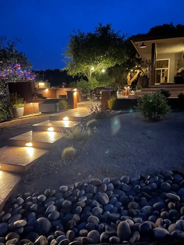 An outdoor patio at night illuminated by pathway lights, with a gravel area in the foreground, potted plants, a tree with string lights, and a house with an open porch in the background.