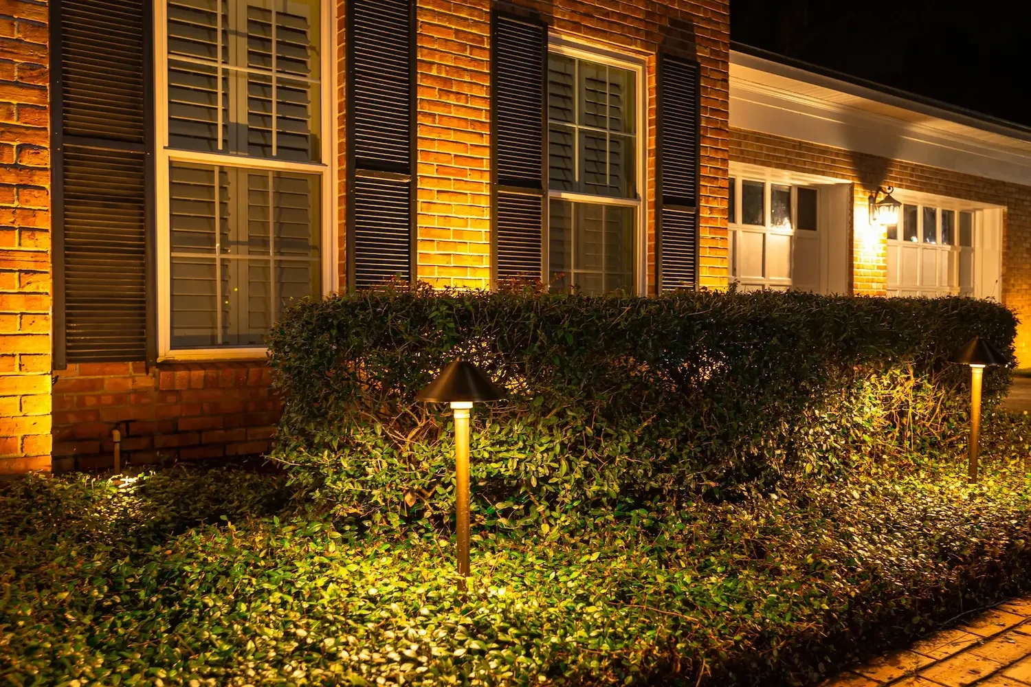 Exterior of a house at night with brick walls, windows with shutters, hedge, and landscape lighting.
