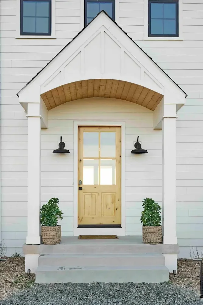 Front porch of a house with a light wooden door, two black wall-mounted lamps, two potted plants on either side, and a small staircase leading to the door.