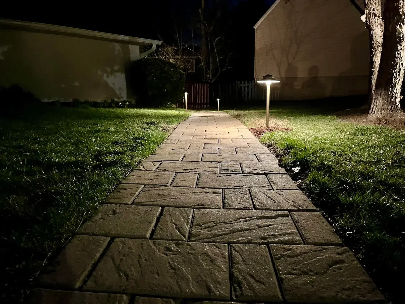Nighttime photograph of a paved pathway illuminated by small landscape lights, with grass on both sides and a house and trees in the background.
