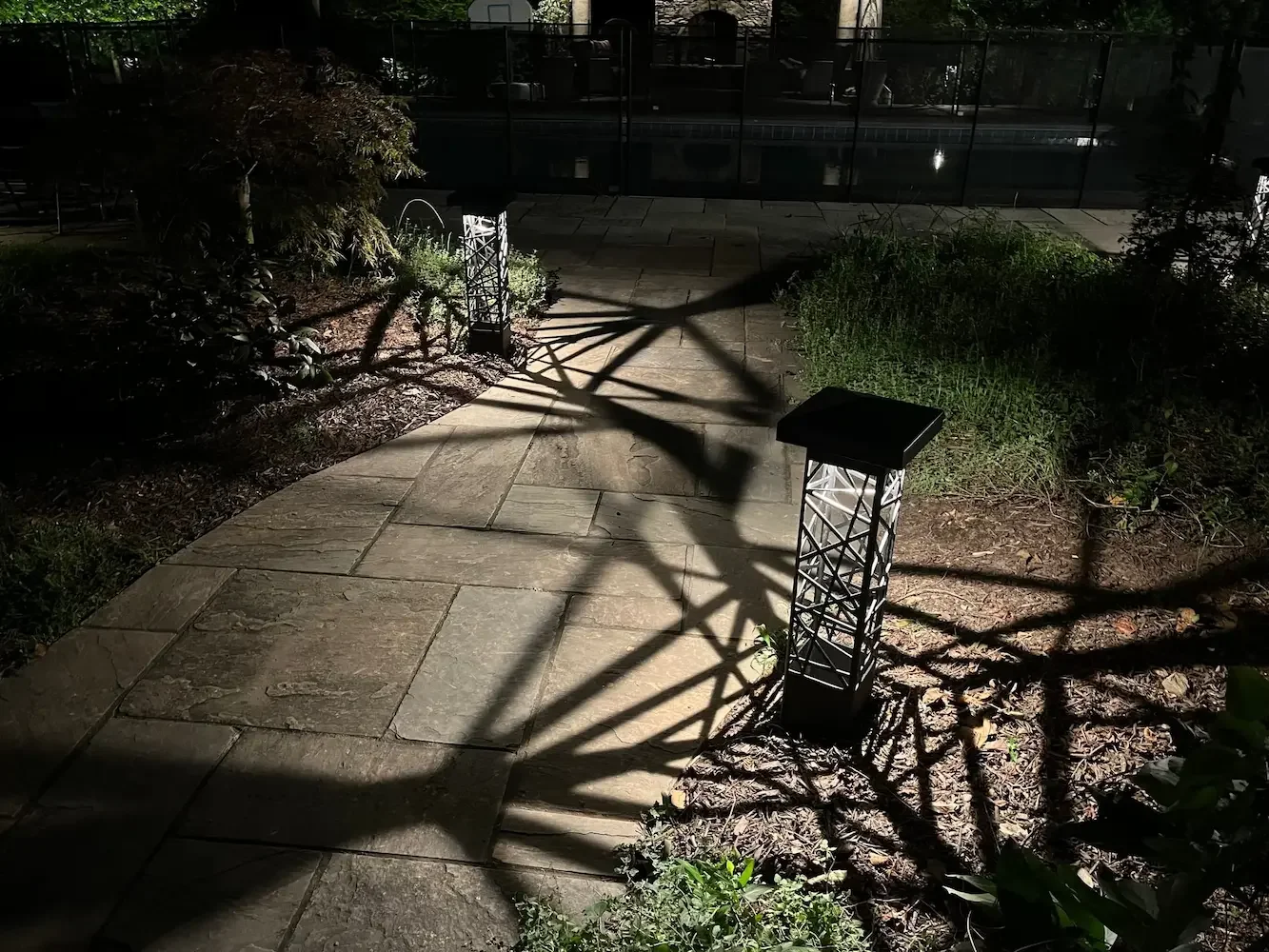 Nighttime outdoor scene showing a stone patio, garden plants, and two decorative lanterns casting shadows on the ground.