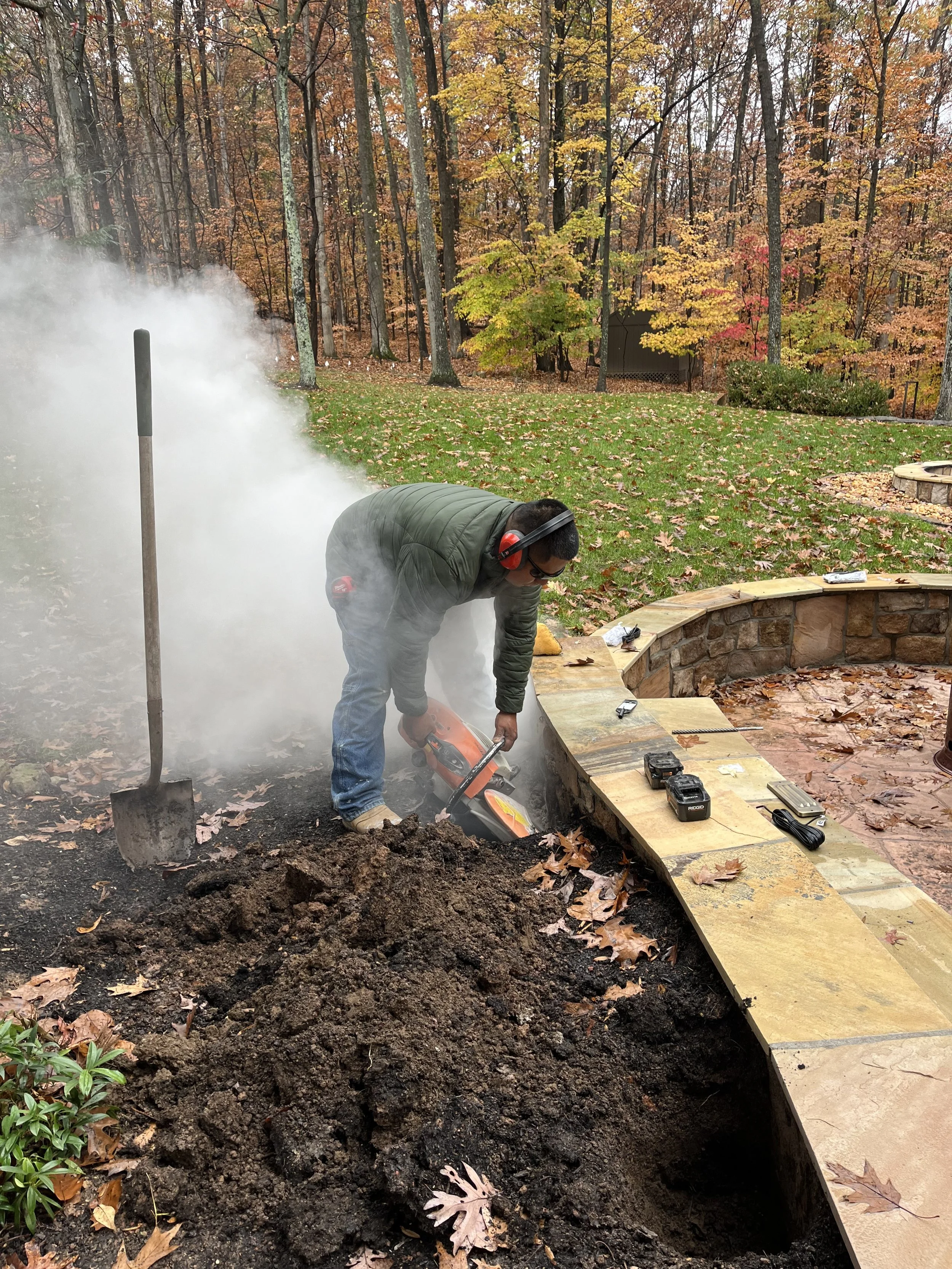 A man wearing a green jacket, jeans, and headphones is cutting a tree root with a chainsaw in a backyard with a curved stone fire pit, leaves on the ground, and trees with autumn-colored leaves in the background.
