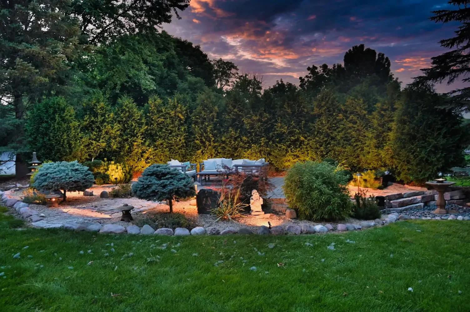 A landscaped backyard garden at dusk with a stone patio, outdoor seating, illuminated pathway, well-maintained grass, and a background of tall trees and a colorful sky.