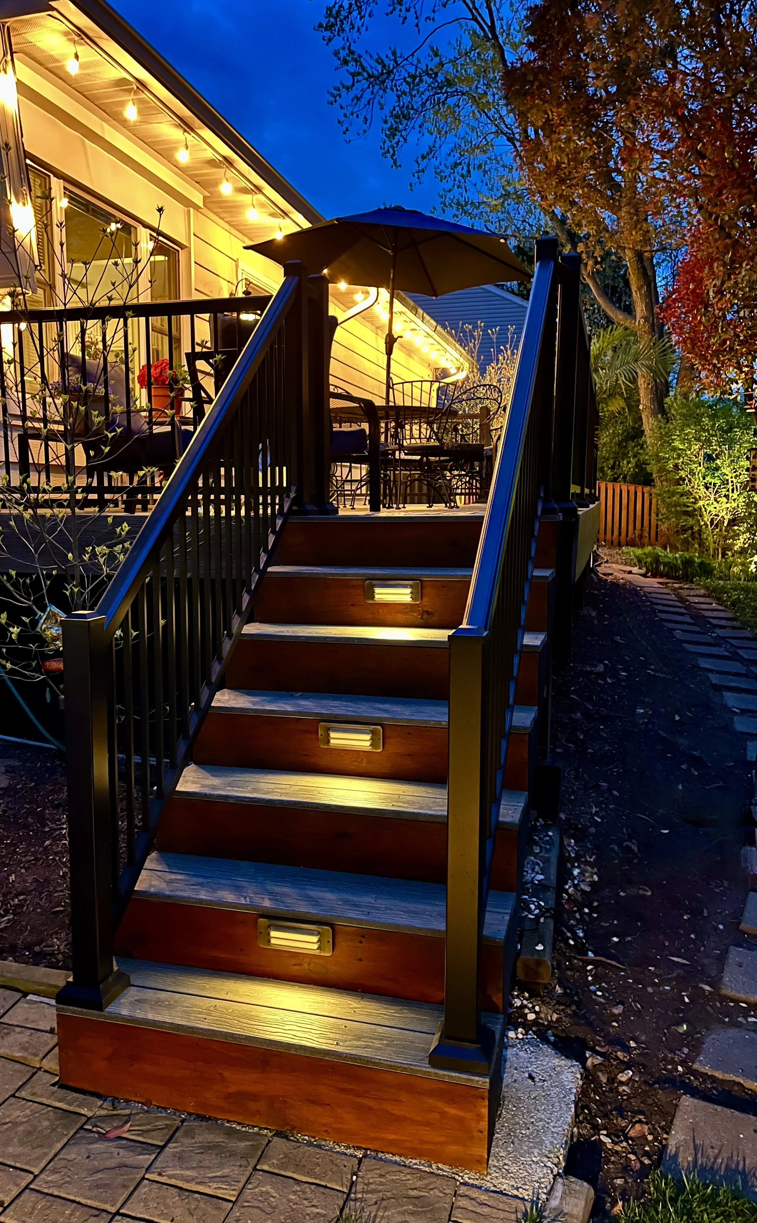 Wooden outdoor staircase with built-in lights leading to a backyard patio with string lights, chairs, table, and an umbrella in the evening. House with lights and trees in the background.