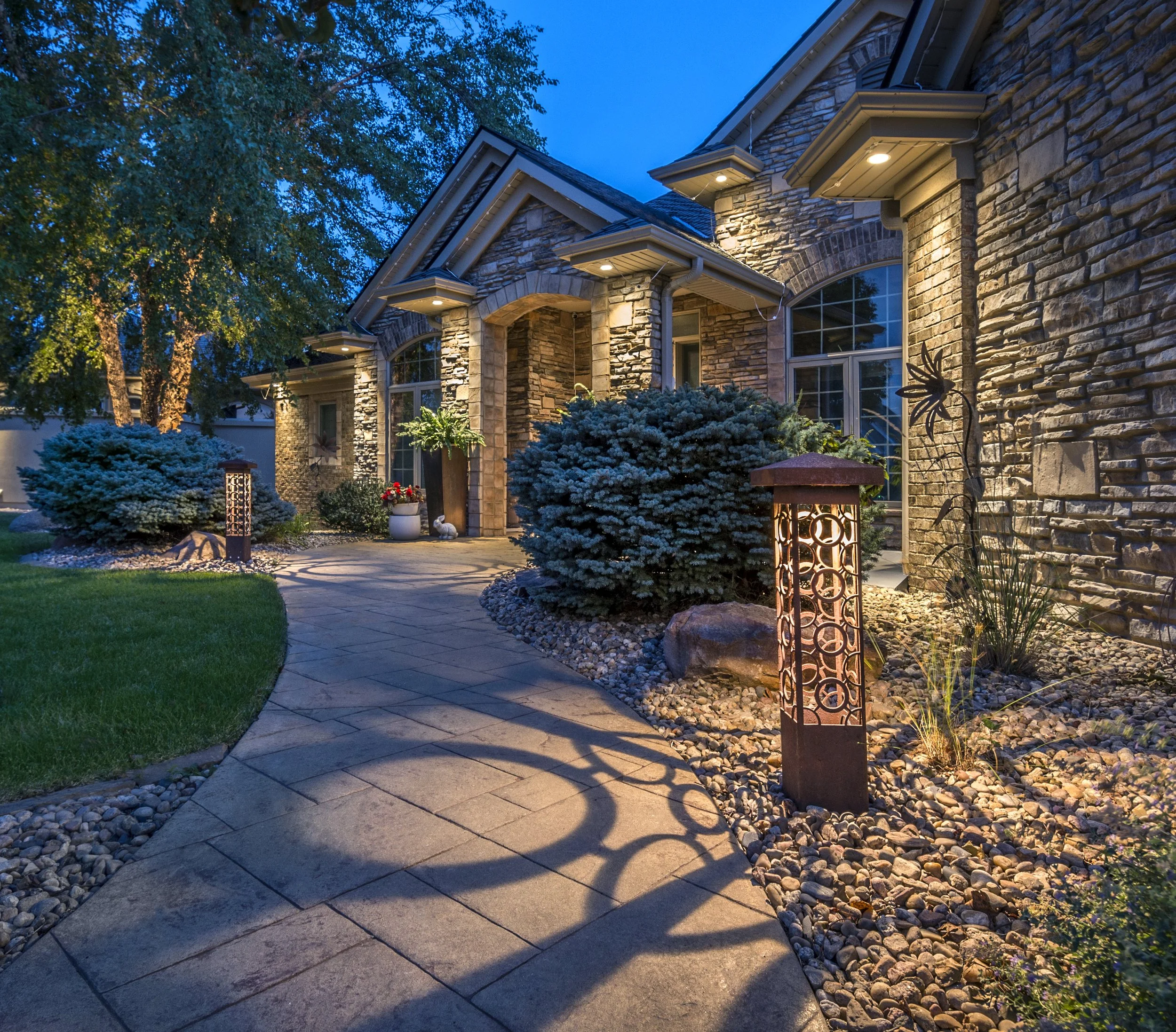 Front yard of a house with stone exterior, illuminated by outdoor lights, with a curved pathway, bushes, potted plants, and decorative outdoor lighting at dusk.