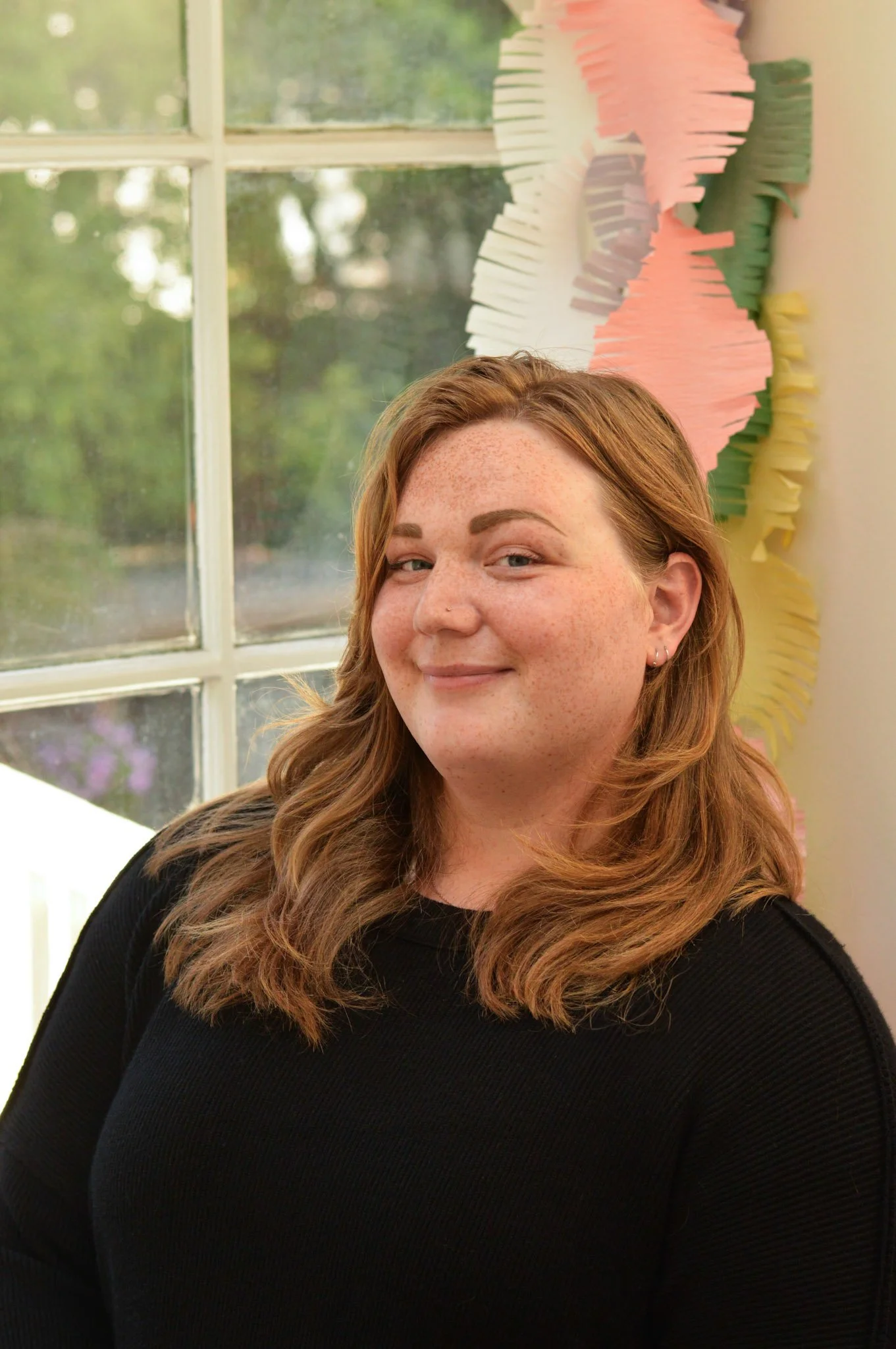 A red haired woman wearing a black top and smiling warmly is sitting in front of a window, with trees outside and colorful paper decor inside