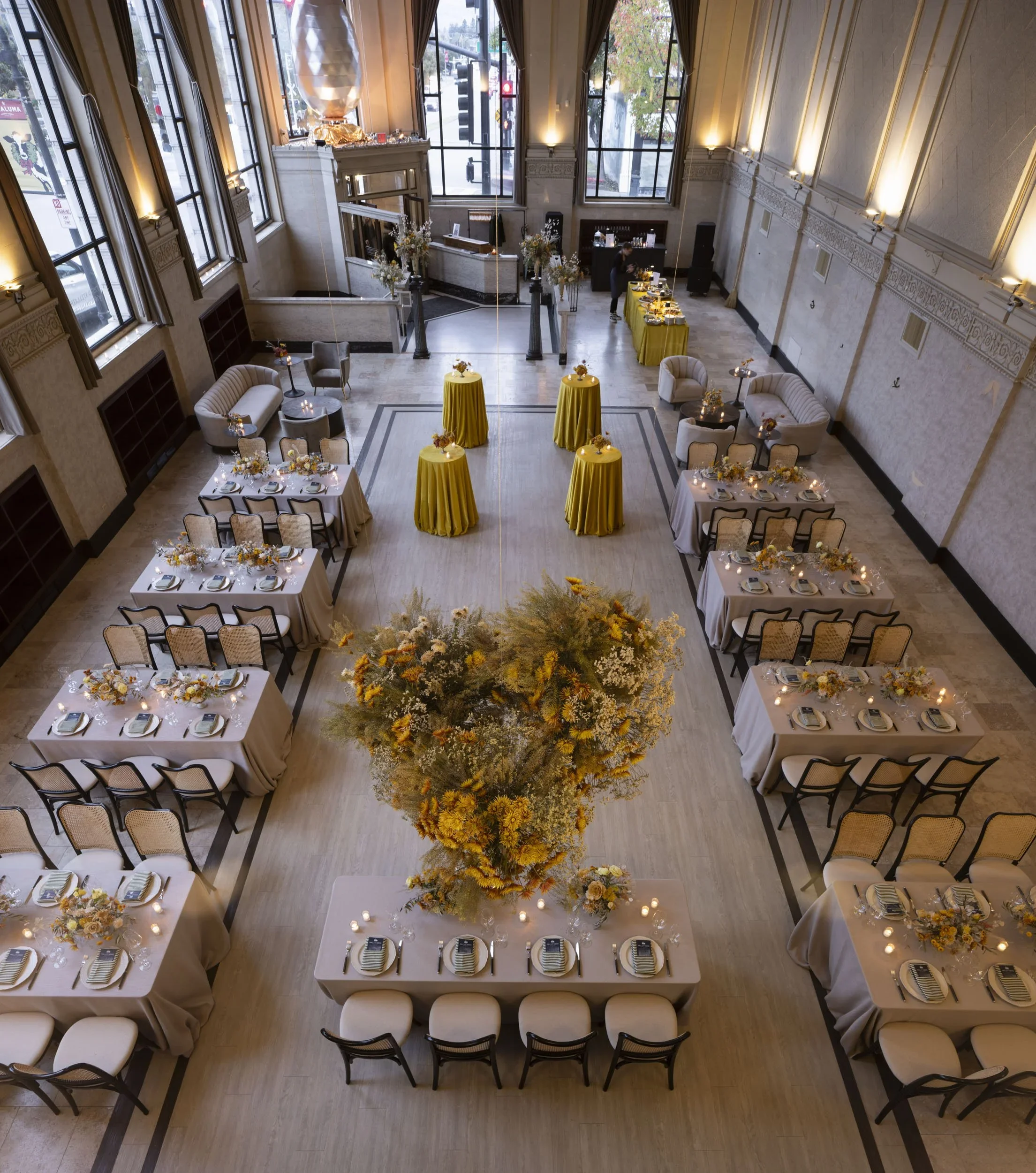 An elegant indoor event space with high ceilings and large windows. The room is decorated with floral arrangements and set for a formal gathering, featuring round tables with floral centerpieces, place settings, and beige upholstered chairs. Several tall tables with yellow tablecloths and flowers are also present, along with a central large floral display. There is a reception desk near the front with a staff member working behind it and a buffet table on the side. The lighting is warm and inviting.