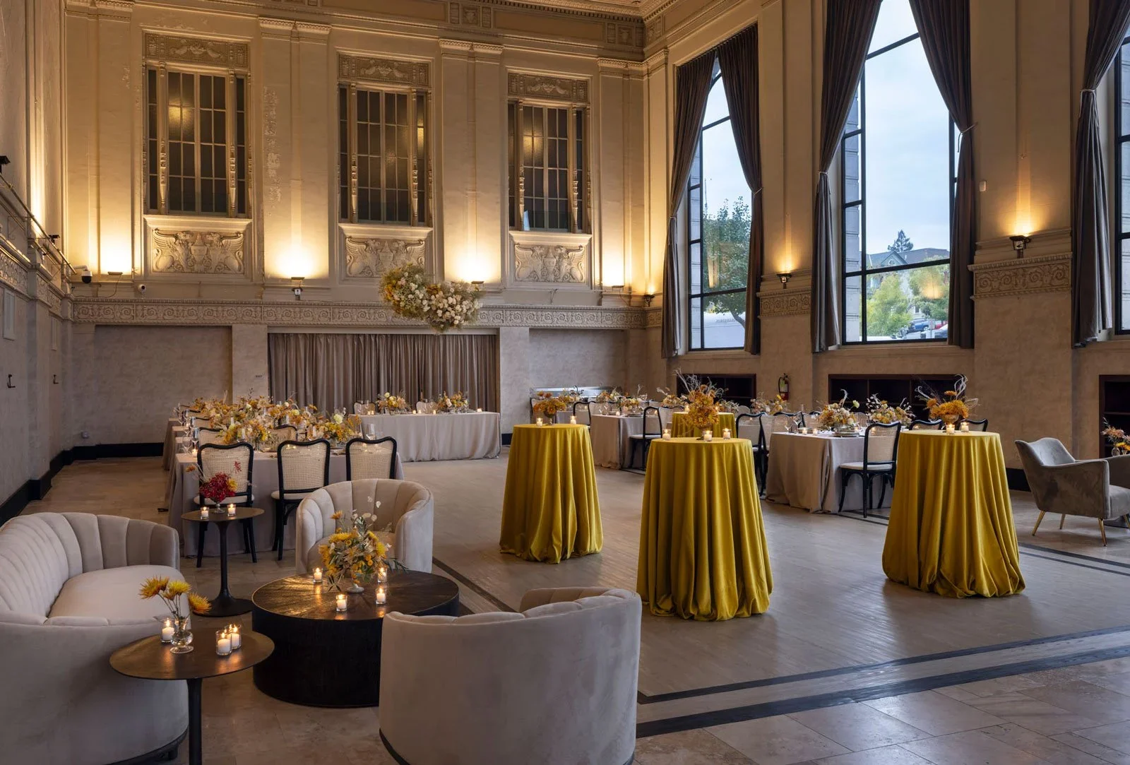 Elegant banquet hall decorated for an event with round tables covered in yellow tablecloths, floral centerpieces, couches, and candles, large windows with dark curtains, and a high ornate ceiling with wall sconces.
