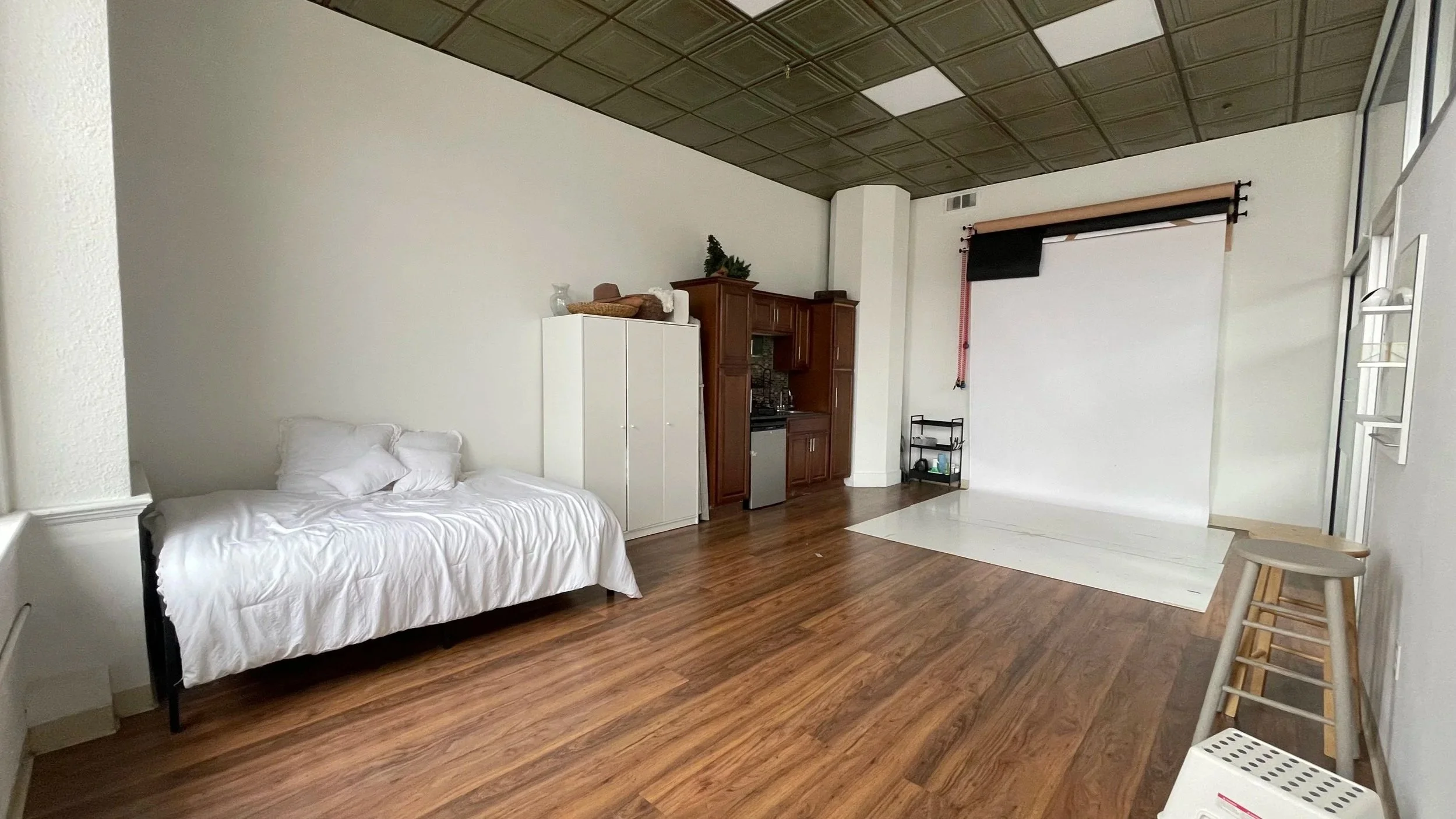 Empty room with hardwood floor, white walls, a white bed with pillows, a mini kitchen with brown cabinets, a white wardrobe, and a photo backdrop setup with lighting equipment.