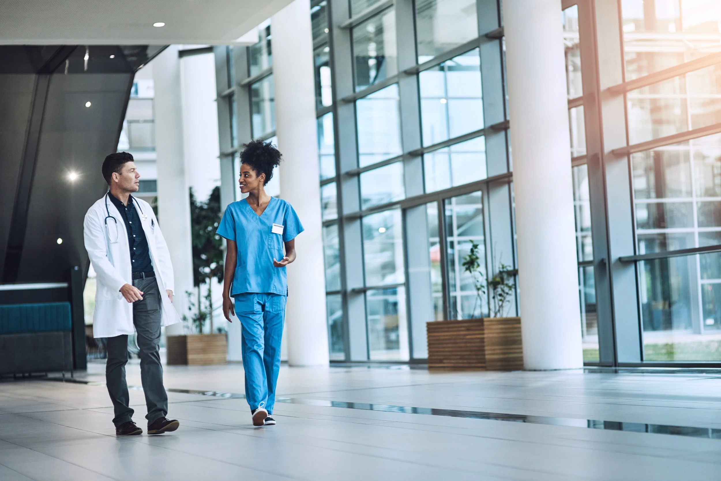 A male doctor and a female nurse are walking together inside a modern hospital lobby with large glass windows and natural sunlight.