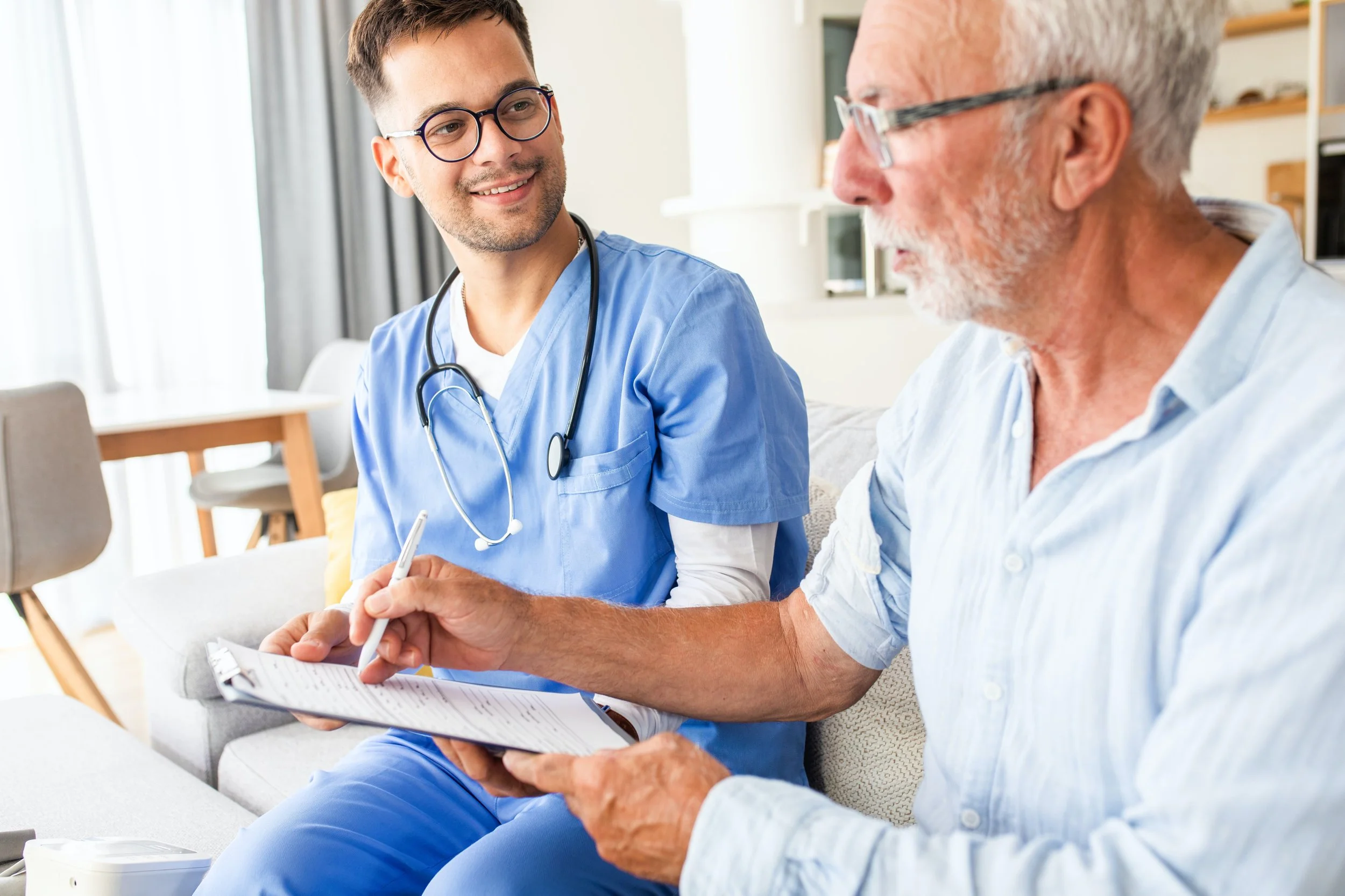 A young male healthcare professional in blue scrubs and glasses talking to an older male patient with white hair and glasses, who is sitting on a sofa, holding a notepad and pen, in a bright, modern room.