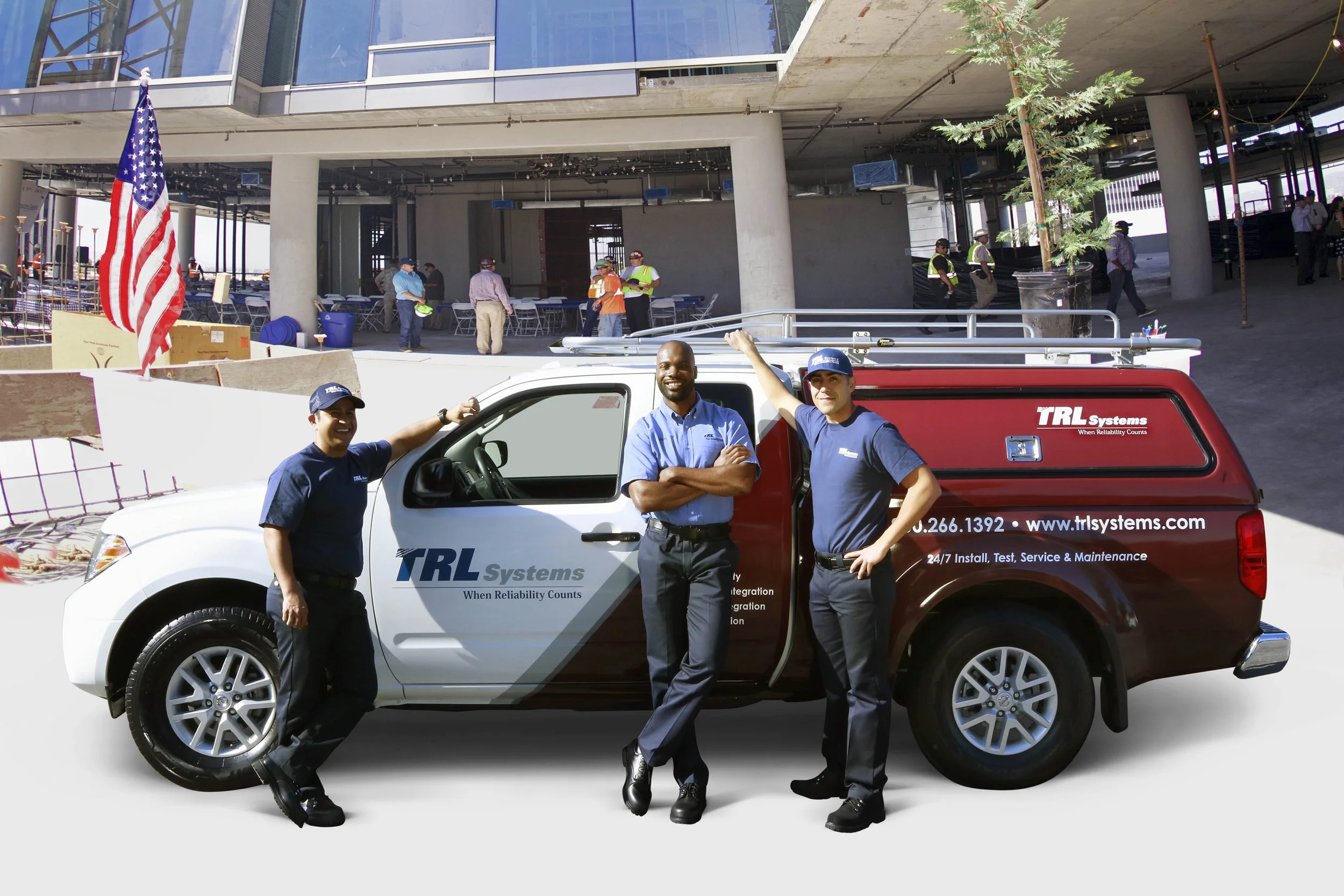 Three men standing in front of a white pickup truck with TRL Systems logo, two men are leaning on the truck, one with arms crossed, wearing dark blue shirts and caps, smiling, in an outdoor construction area with people and construction site in the background.