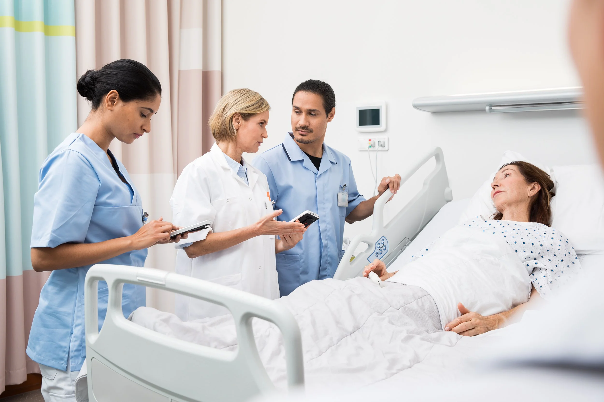 Four medical professionals, three women and one man, are standing around a female patient lying in a hospital bed. The patient appears to be conscious and is wearing a hospital gown, while the medical staff are looking at her and discussing her condi