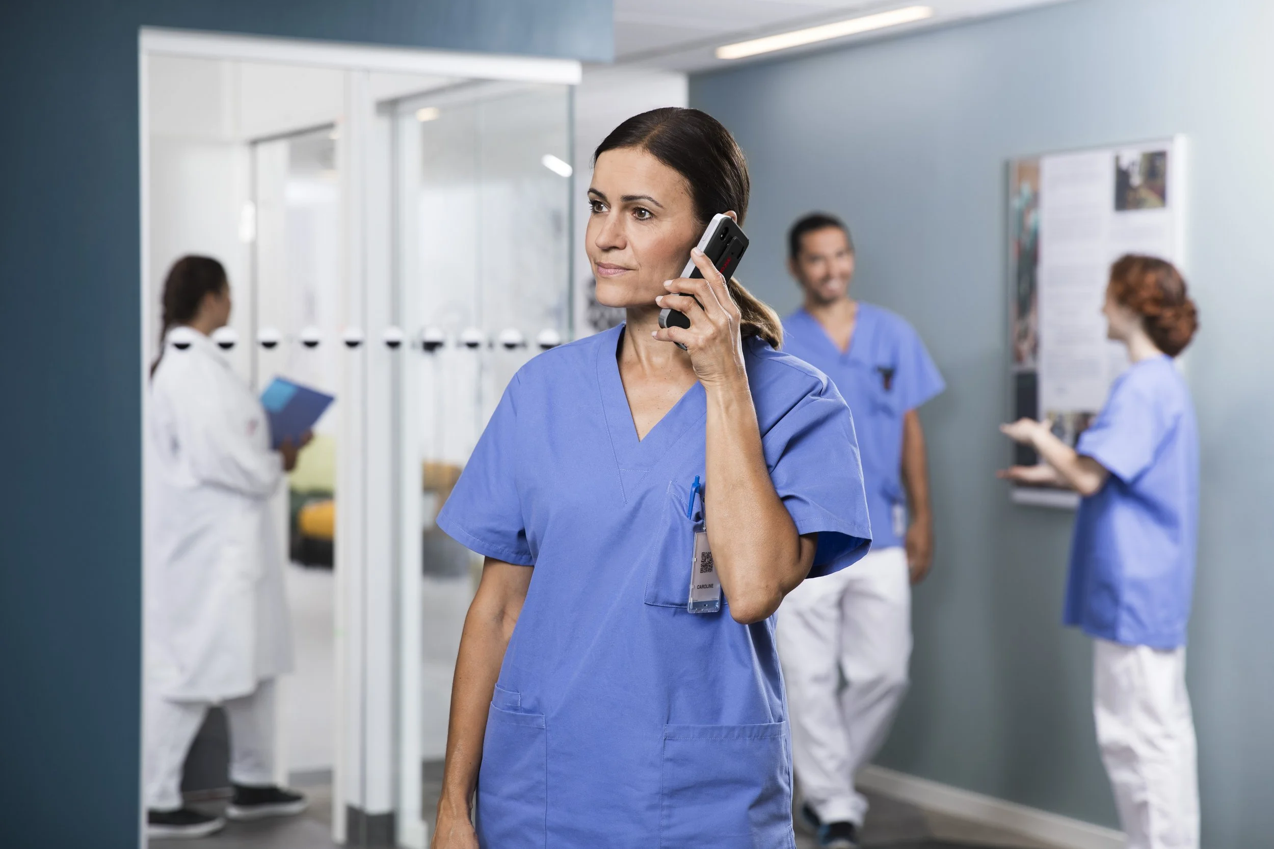 A woman in blue scrubs talking on a cell phone while standing in a medical facility with other healthcare workers in the background.