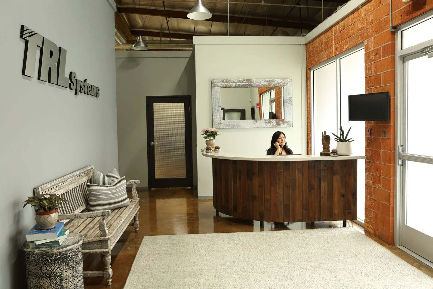Reception area with a woman smiling at a curved wooden desk, large window on the right, brick wall, mirror, and potted plants.