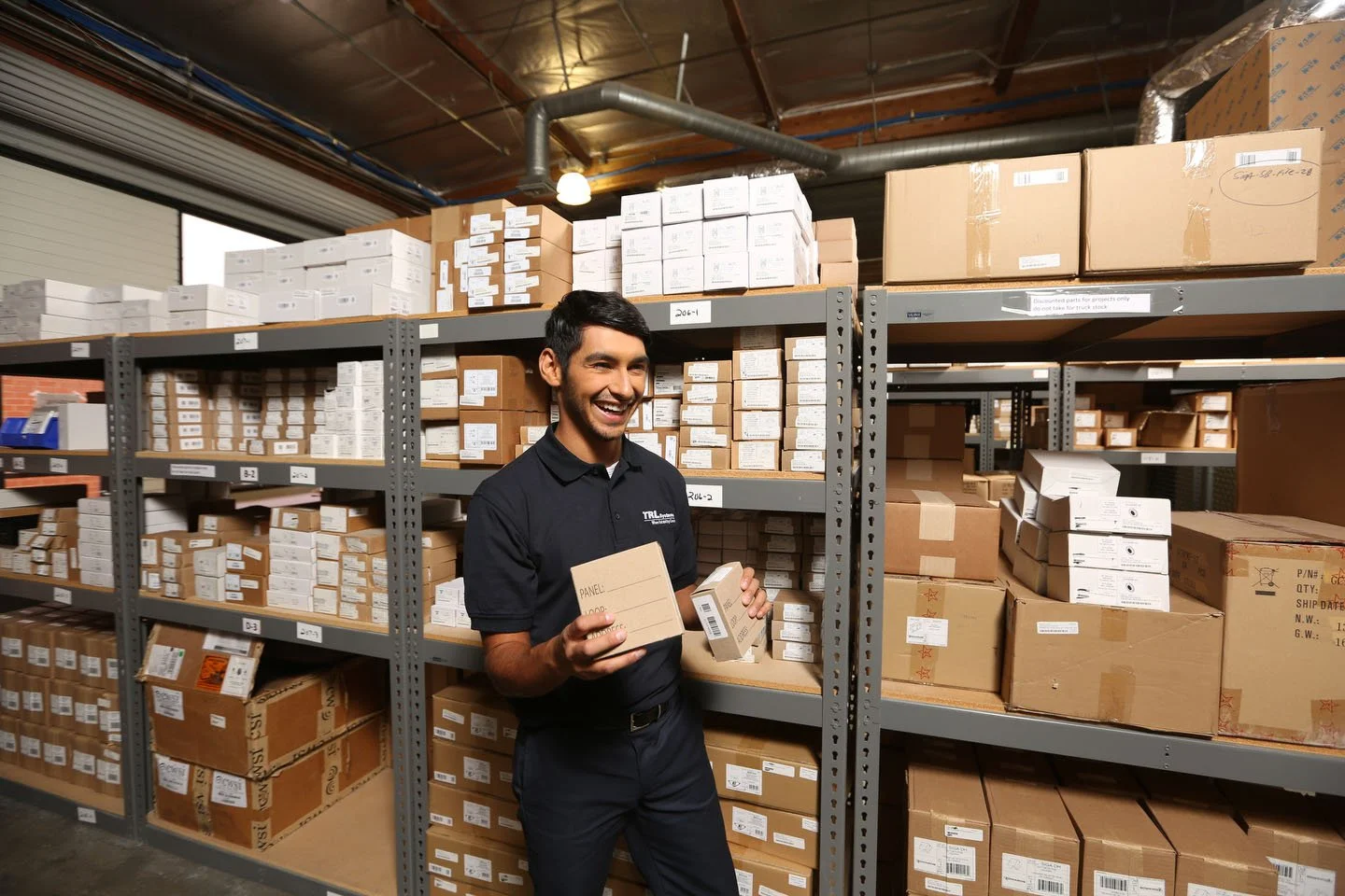 A man smiling and holding two small cardboard boxes in a warehouse aisle with shelves filled with packed boxes and packages.