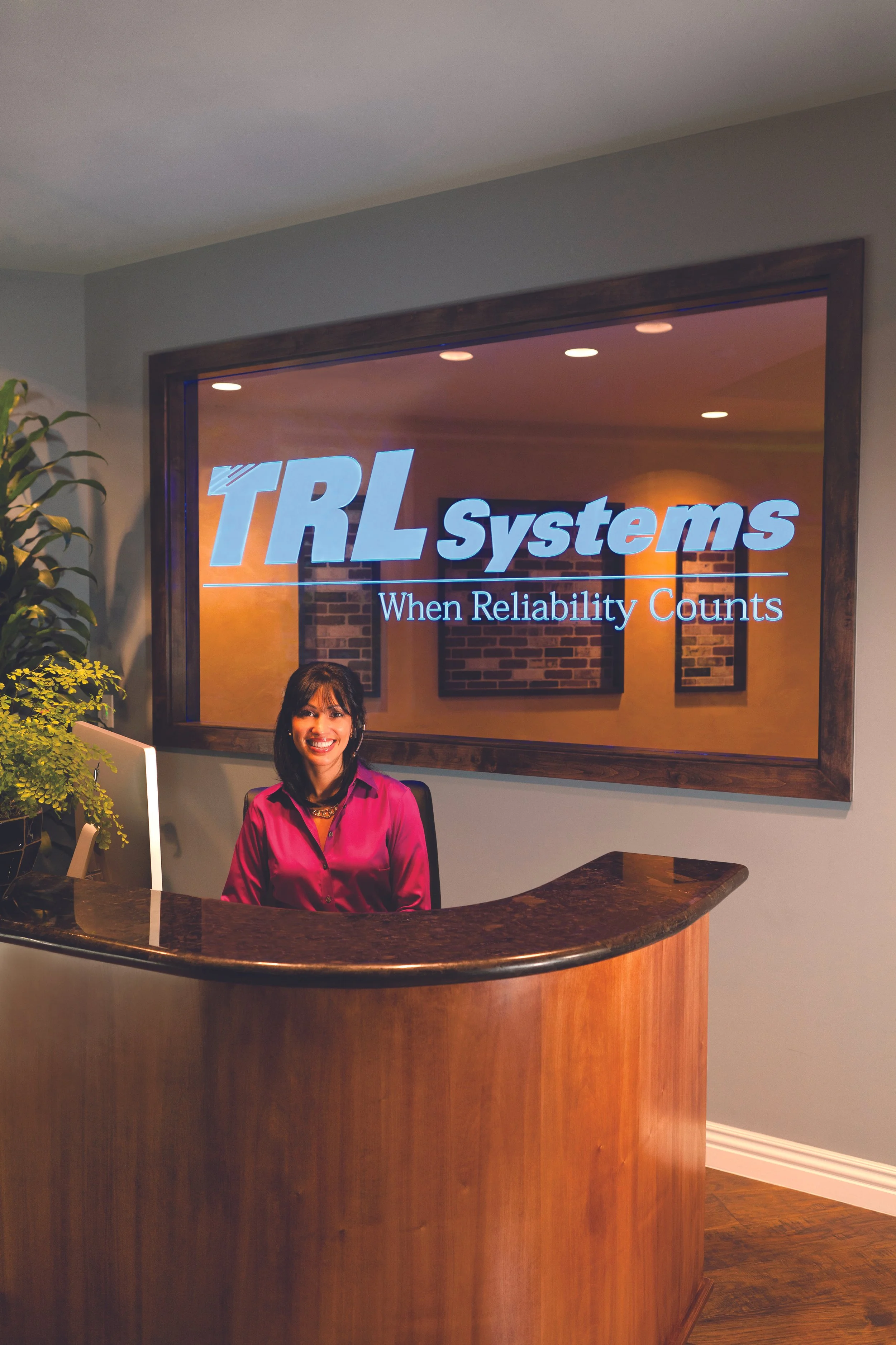 Receptionist sitting at a curved wooden desk with a computer, smiling, in front of a wall with a sign that says "TRL Systems" and the slogan "When Reliability Counts." There are plants on the left and a brick wall background visible through a window.