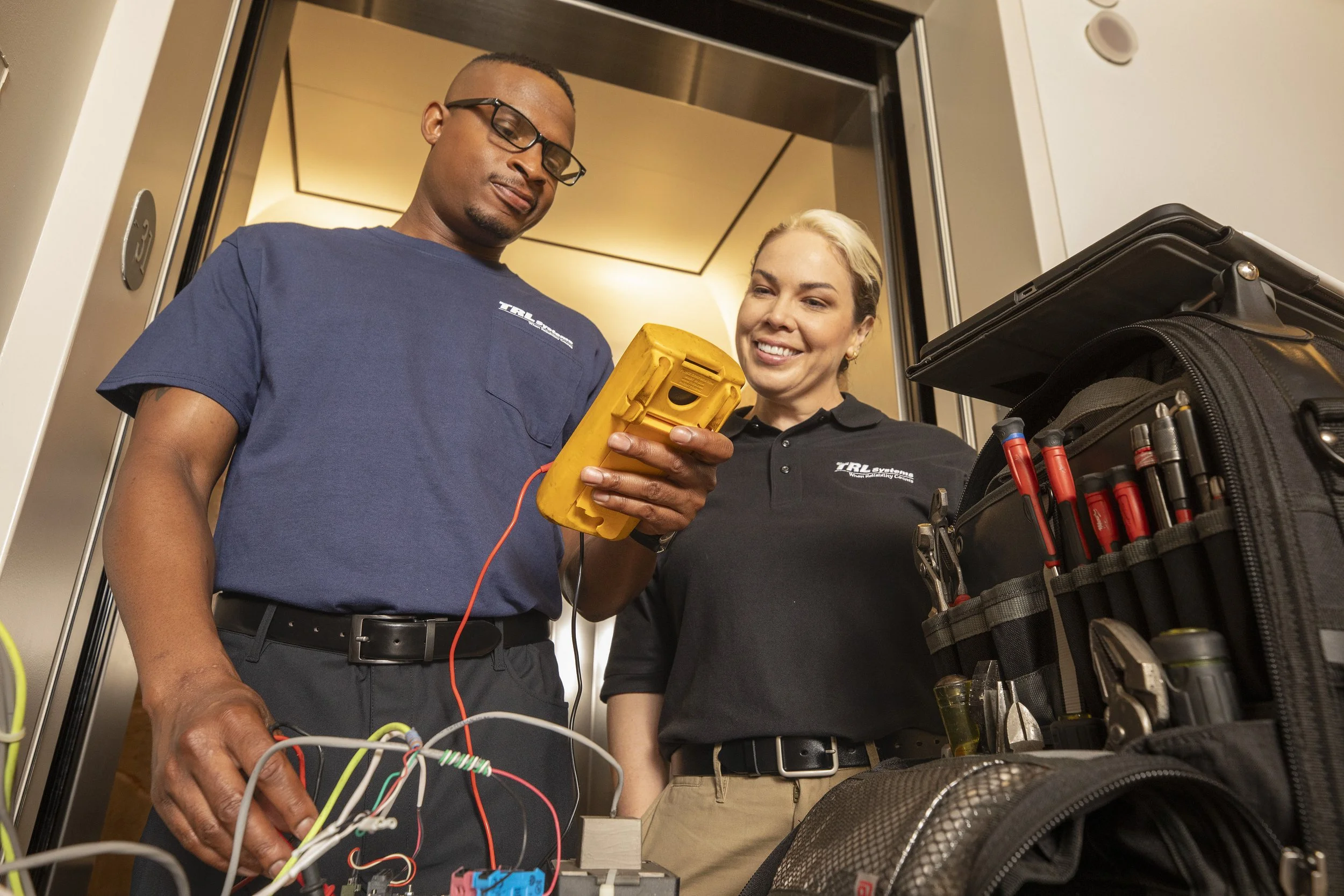 Two technicians working together inside an elevator shaft, one holding a multimeter and the other operating equipment, with tools and a toolbox nearby.