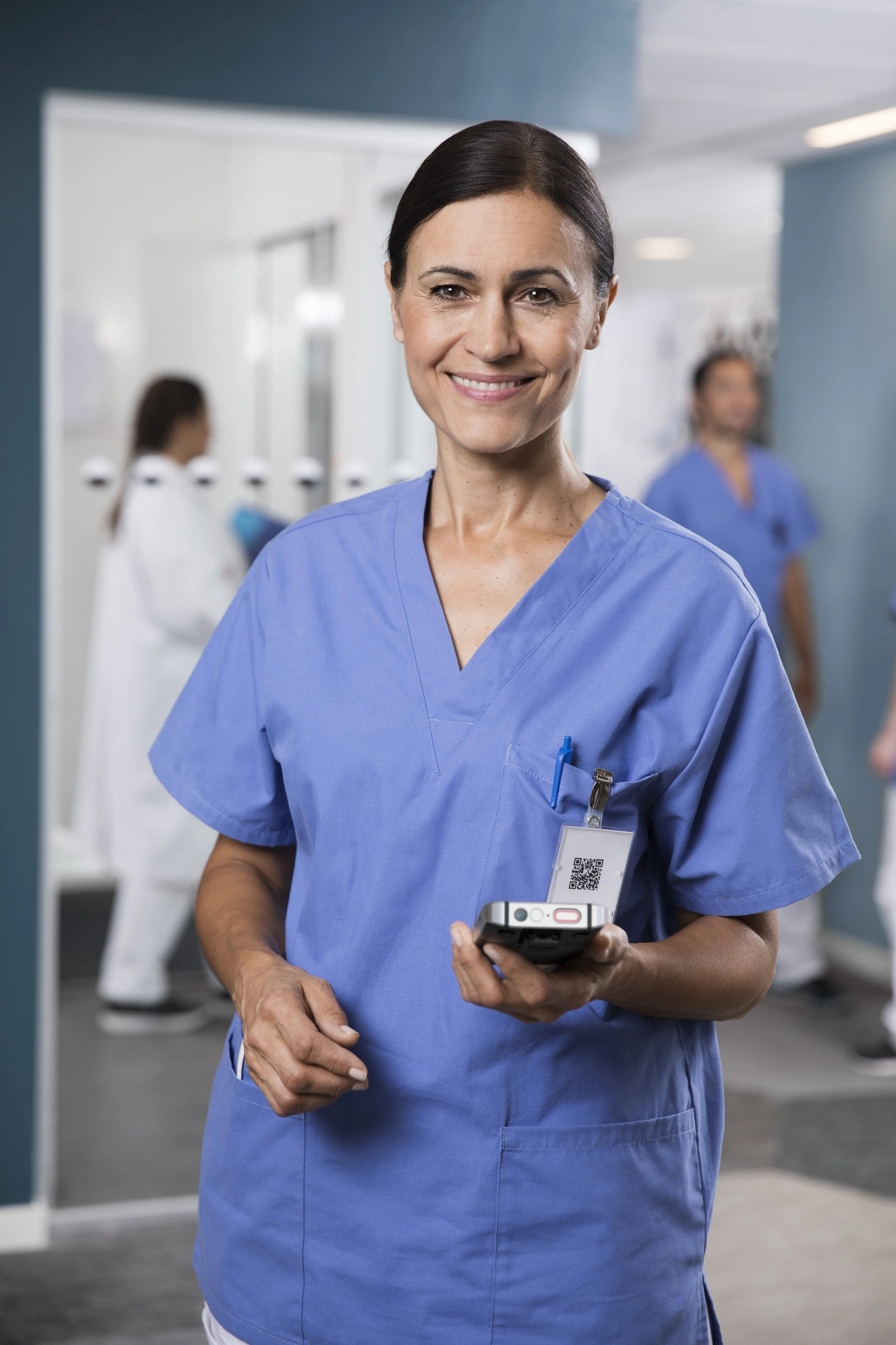 A female healthcare worker in blue scrubs holds a mobile device, standing in a hospital corridor with other healthcare staff in the background.