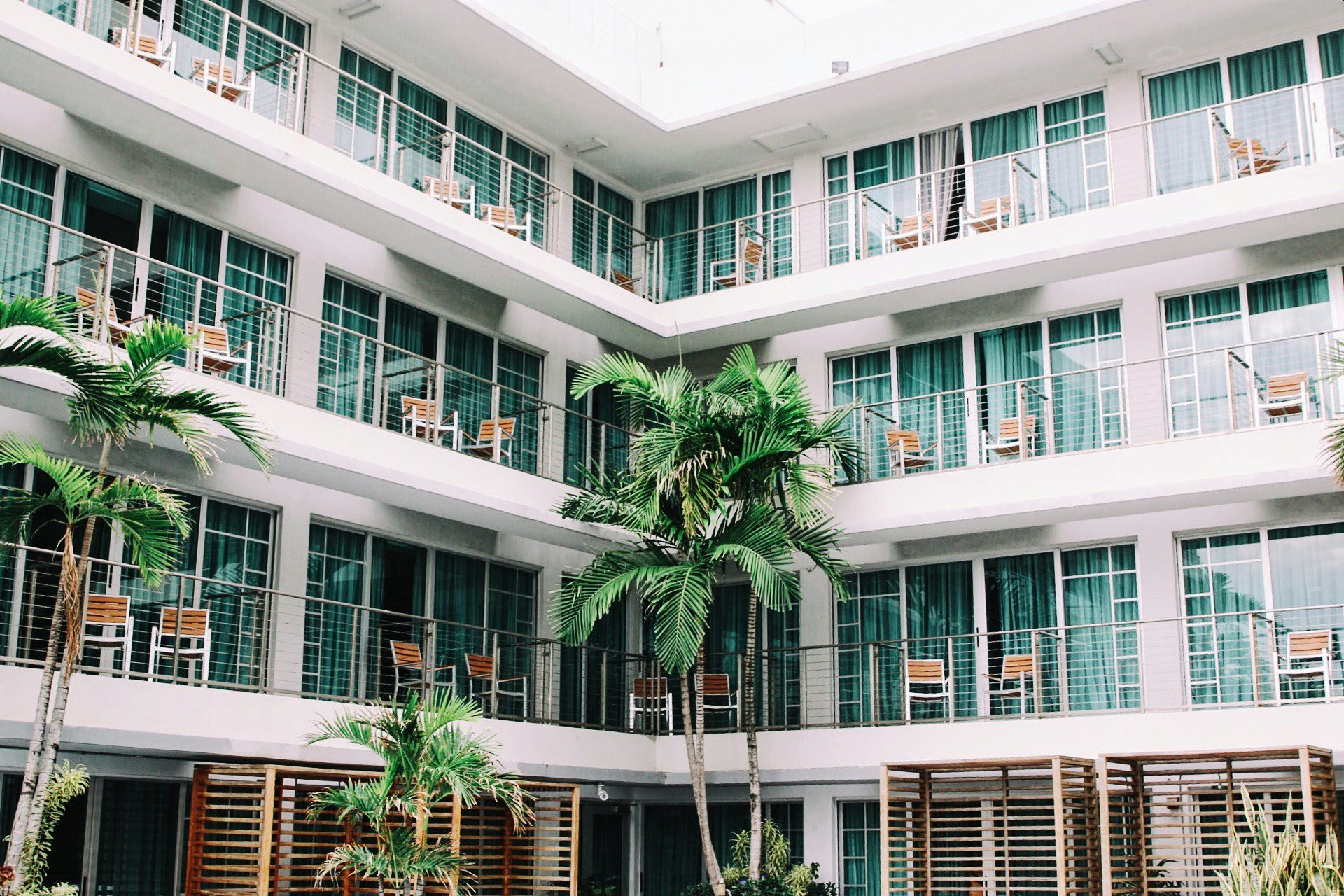 A modern multi-story hotel with white exterior walls, glass sliding doors with teal curtains, balconies with chairs and tables, and landscaped with tall palm trees and lush greenery.