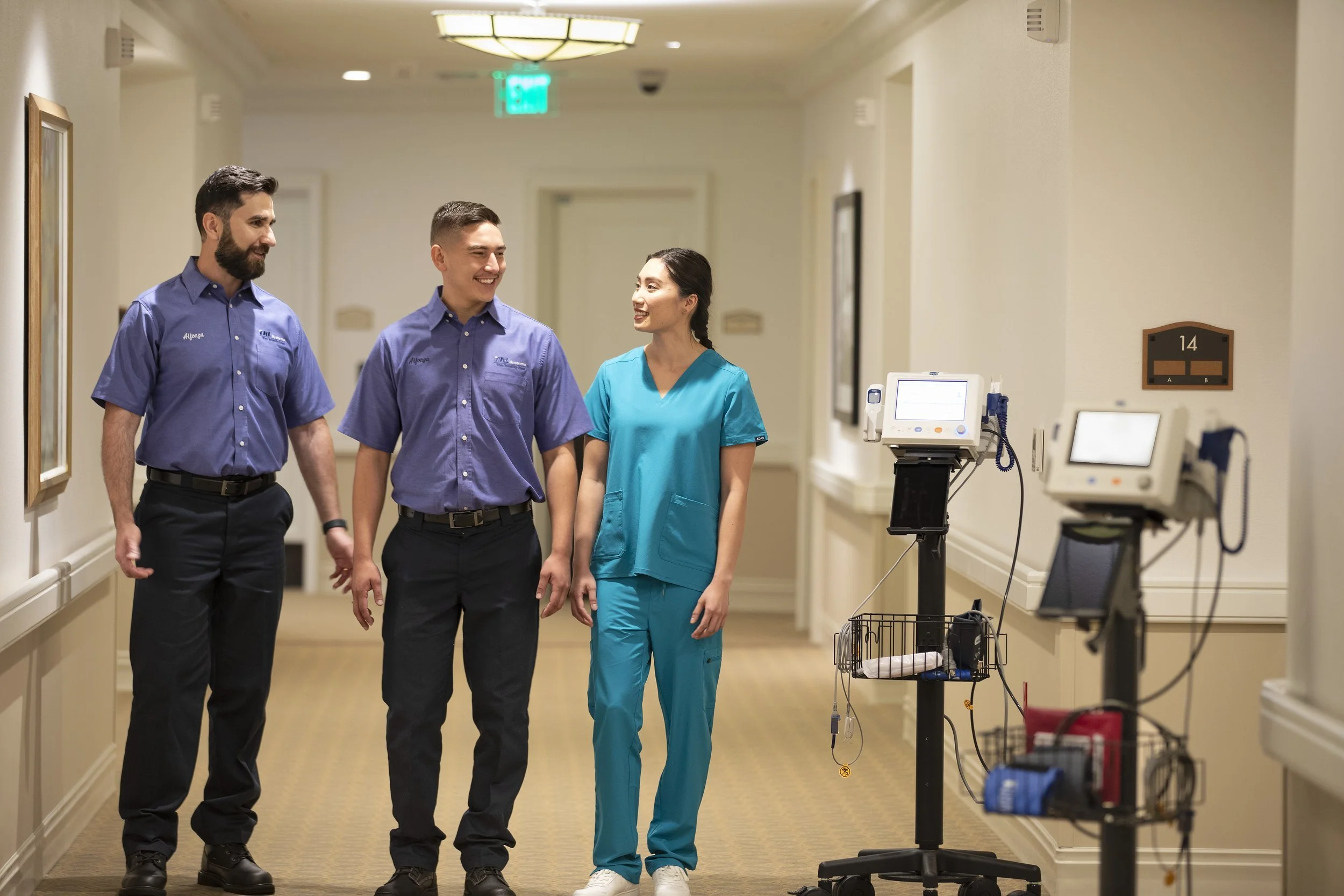 Three medical professionals, two men and a woman, walking down a hospital corridor and smiling at each other. They are wearing scrubs and appear to be engaged in a friendly conversation. Medical equipment is visible on the right side of the image.