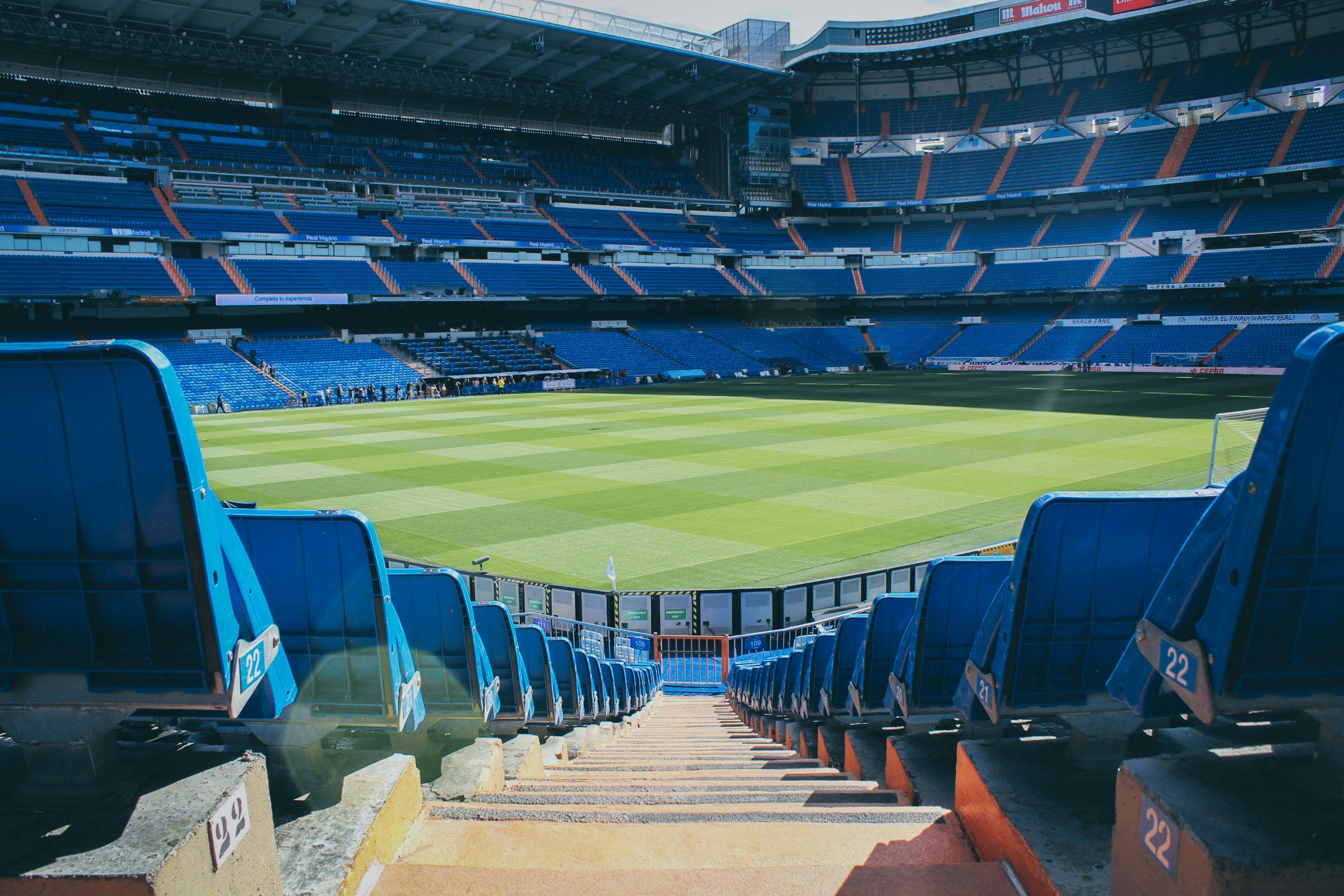 View of a large empty soccer stadium with bright green grass, blue seating, and a few people near the field. The photo is taken from the perspective of someone sitting in the stands, looking toward the field.