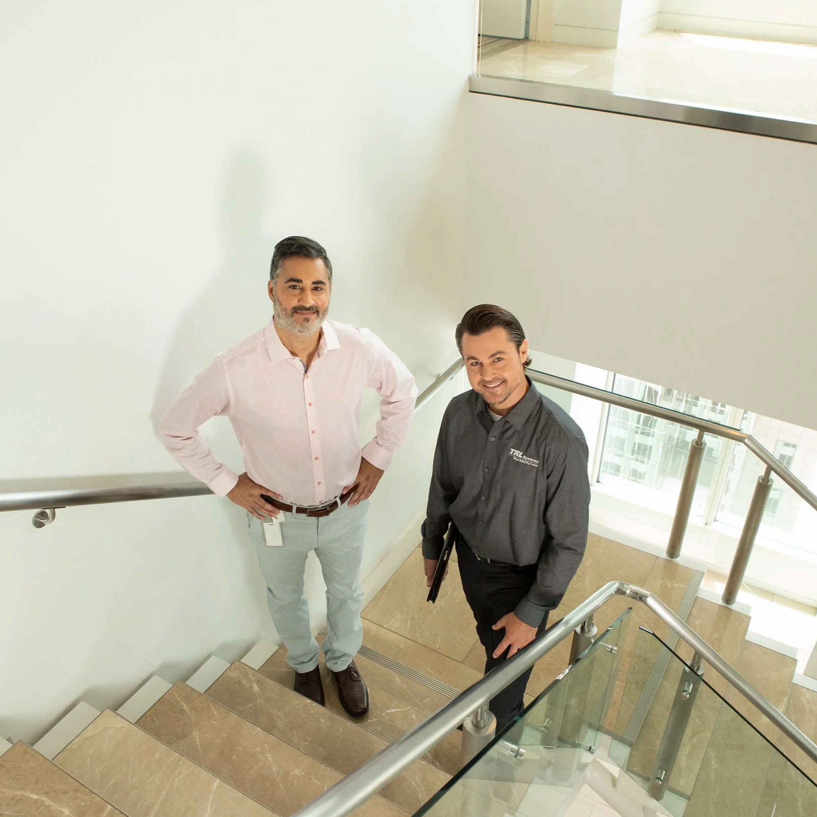Two men standing on a staircase landing in an office building, looking up at the camera. One man is wearing a white button-down shirt and light-colored pants, while the other is in a dark gray shirt with company logos and dark pants. The staircase has a glass and metal railing, and the background includes a large window with a view of a modern building outside.