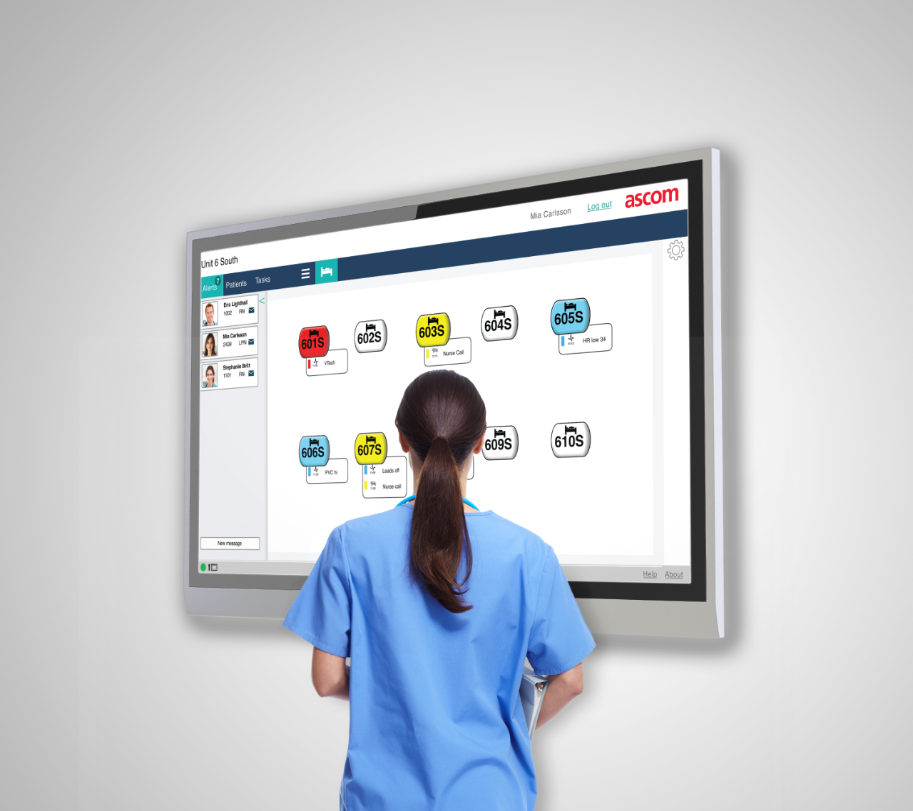 A nurse in blue scrubs standing in front of a large hospital computer screen displaying patient information and task schedules in a medical setting.