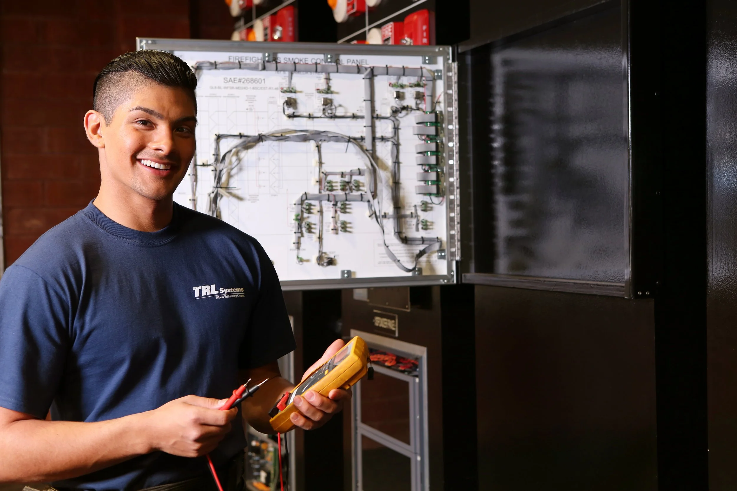 A young man smiling, holding a multimeter with probes in front of an electrical panel or circuit board.