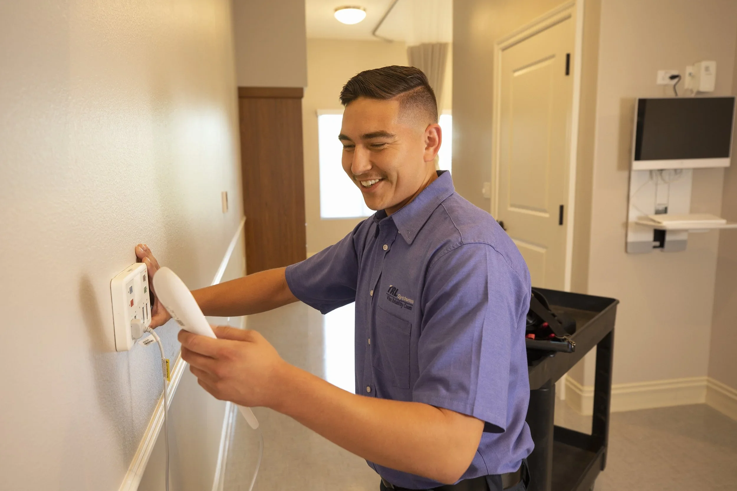 A technician in a purple shirt adjusting a thermostat on a beige wall in a room with a window and various electronic devices.