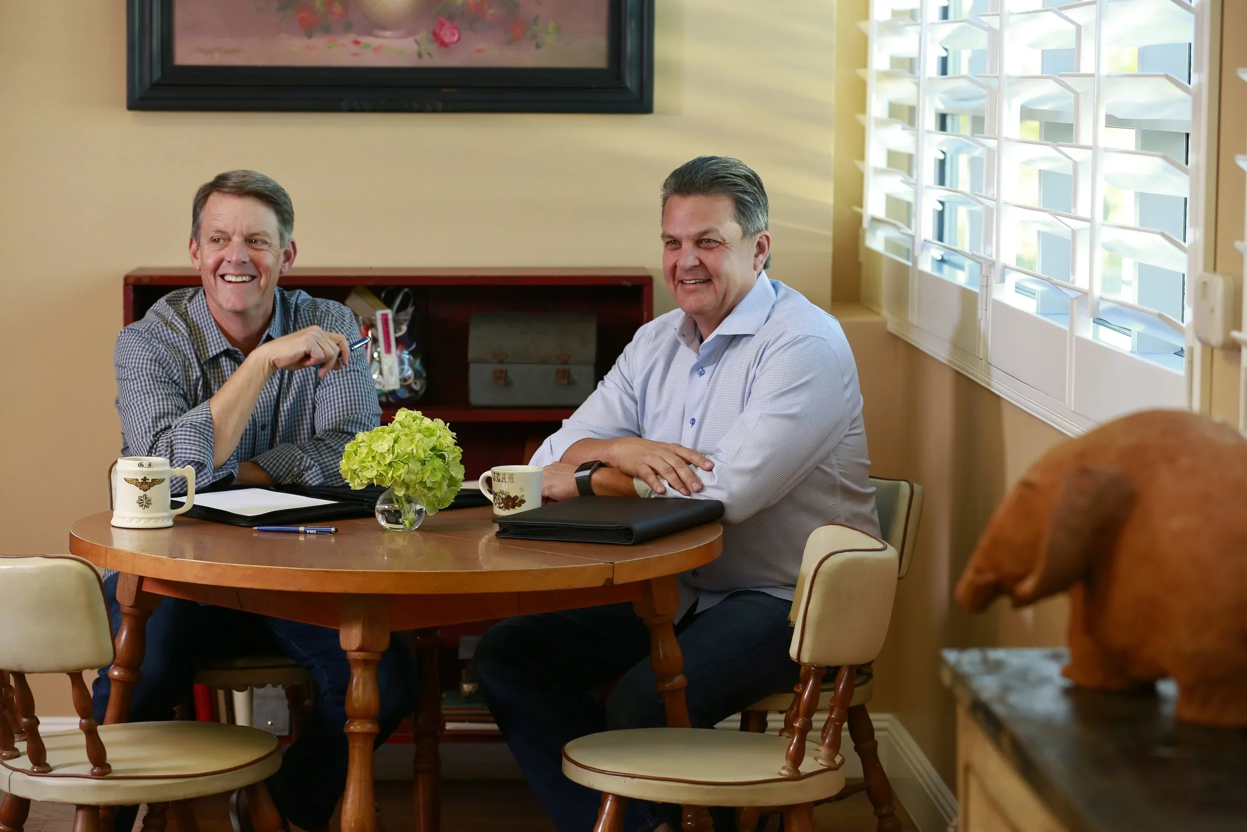 Two middle-aged men sitting at a wooden table in a brightly lit room, smiling, with a vase of green flowers, and two mugs in front of them. One man is wearing a checkered shirt, and the other is in a light blue shirt. There are large windows with white blinds behind them, and a framed painting on the wall.